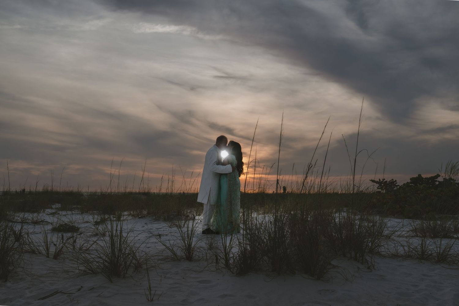 Couple embracing at sunset on a sandy beach with sea oats and dramatic cloudy sky in background.
