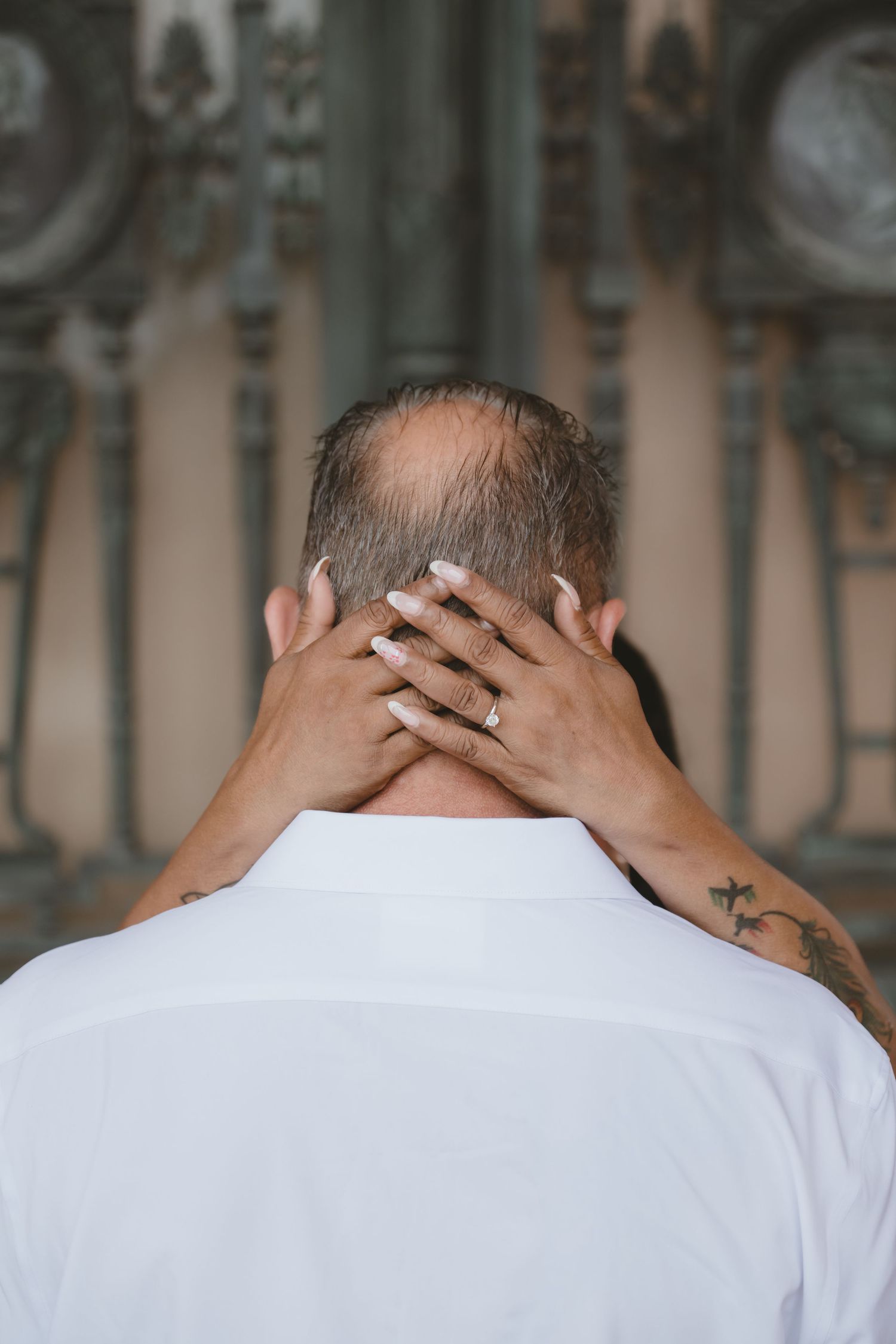 Back view of a man in white shirt with tattooed arms and a woman's hands with ring resting on his neck.