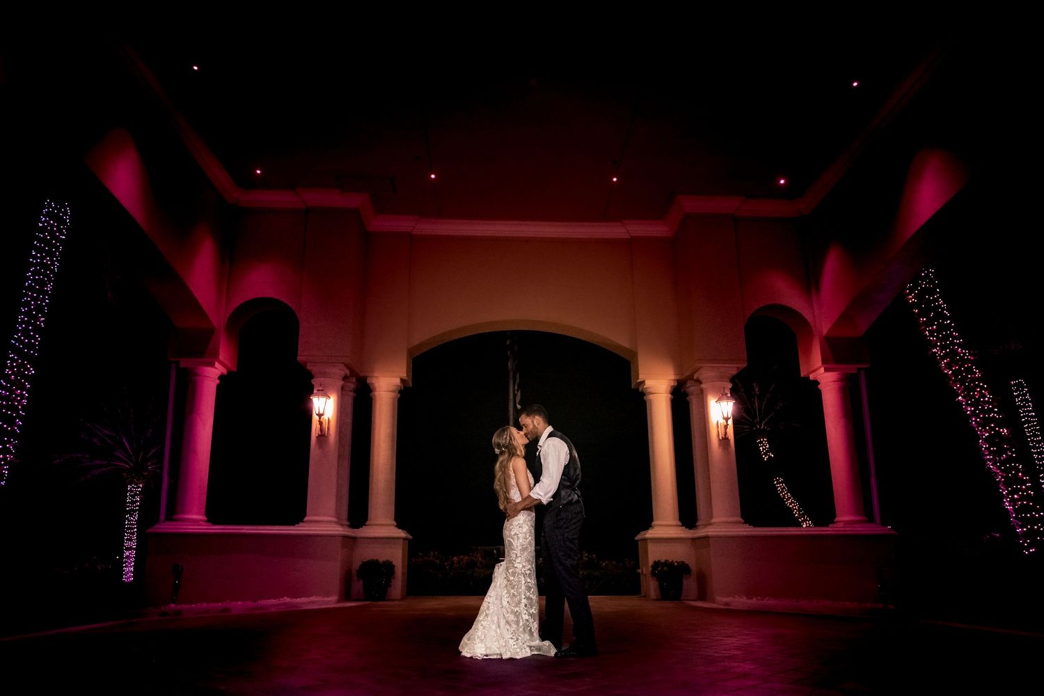Bride and groom share a romantic kiss under dramatic pink uplighting at a stunning wedding venue.