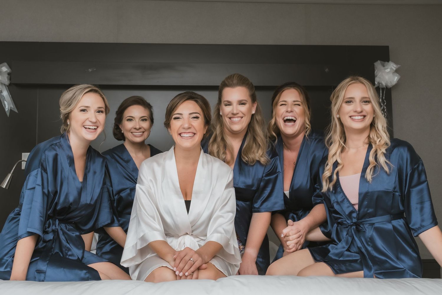 Bride and six bridesmaids in navy satin robes smiling together during wedding morning preparations.
