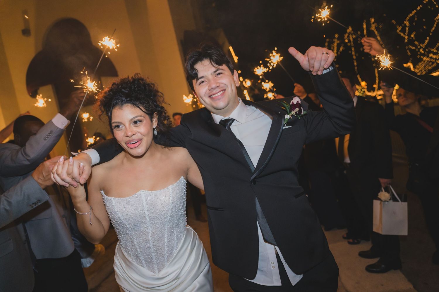 Joyful newlywed couple exits wedding reception through sparkler tunnel, bride in strapless gown, groom in black tuxedo.