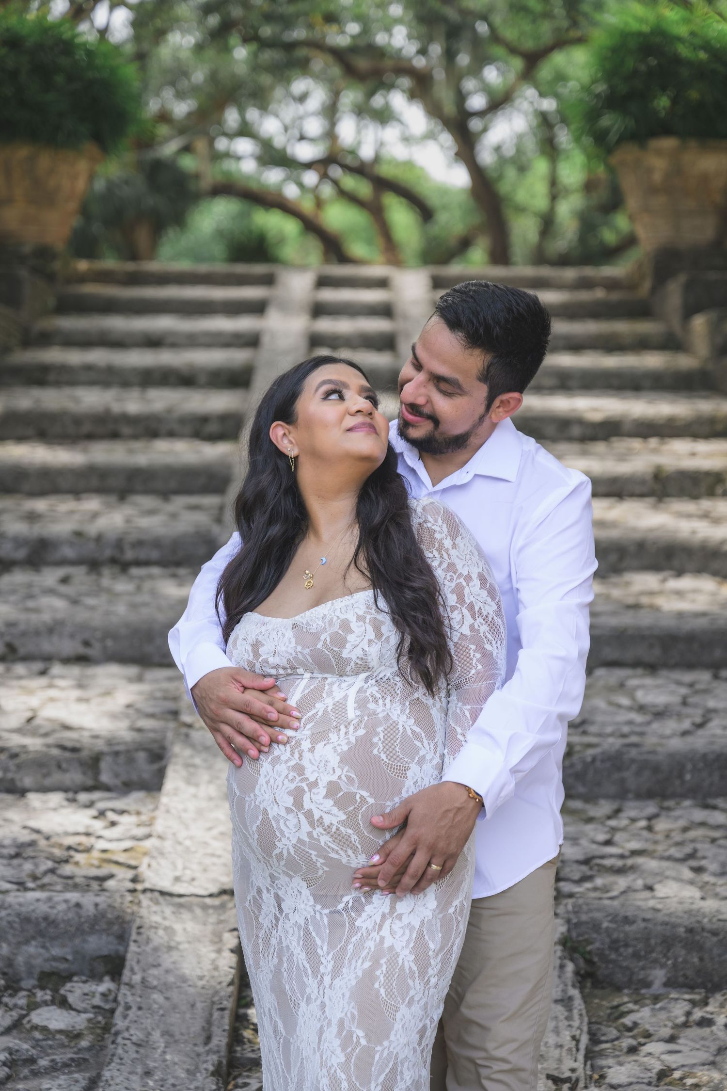 Couple embracing outdoors, pregnant woman in white lace gown, man in white shirt, stone steps and trees in background.
