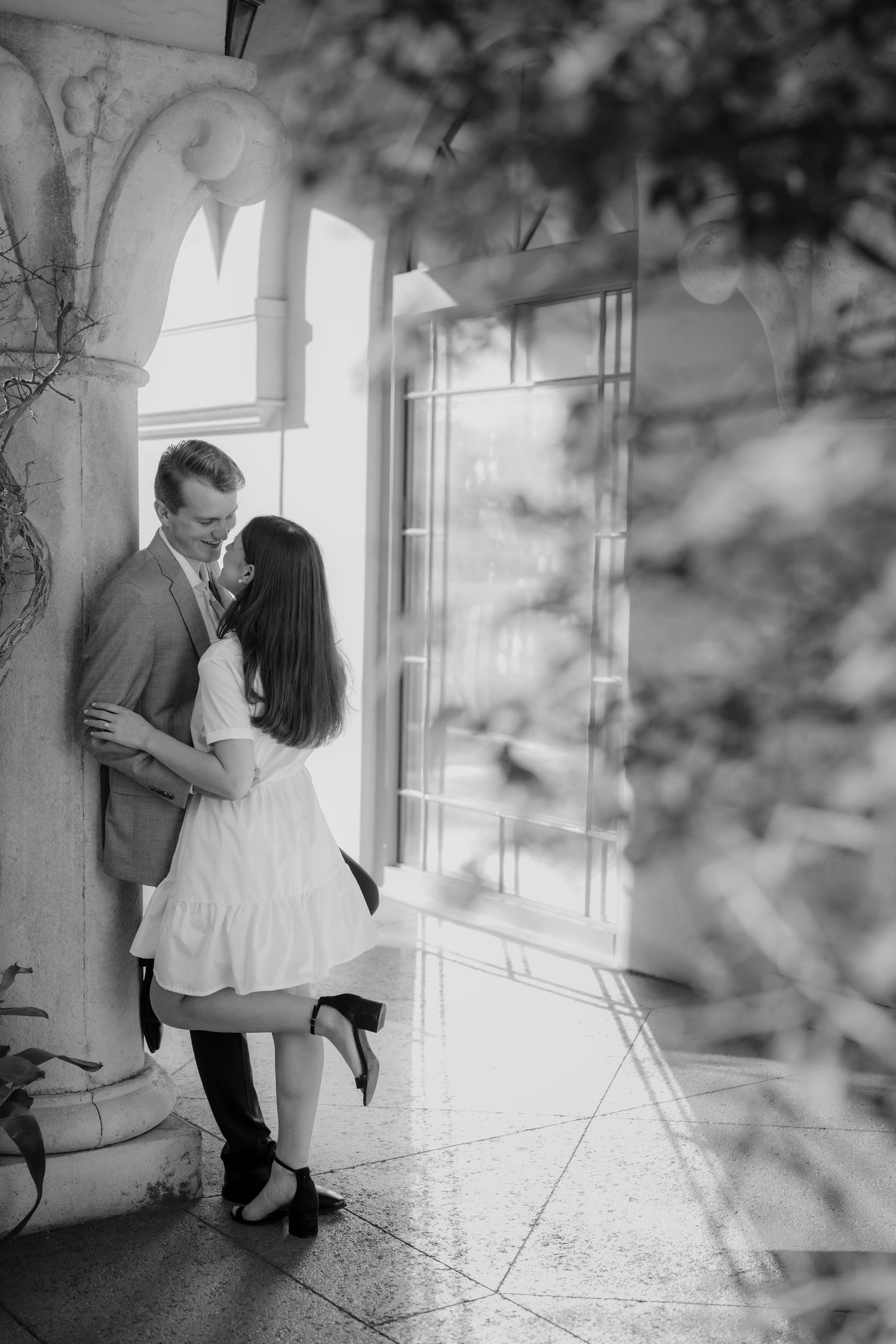 Black and white photo of a couple sharing a kiss beside a ornate column near a bright window.