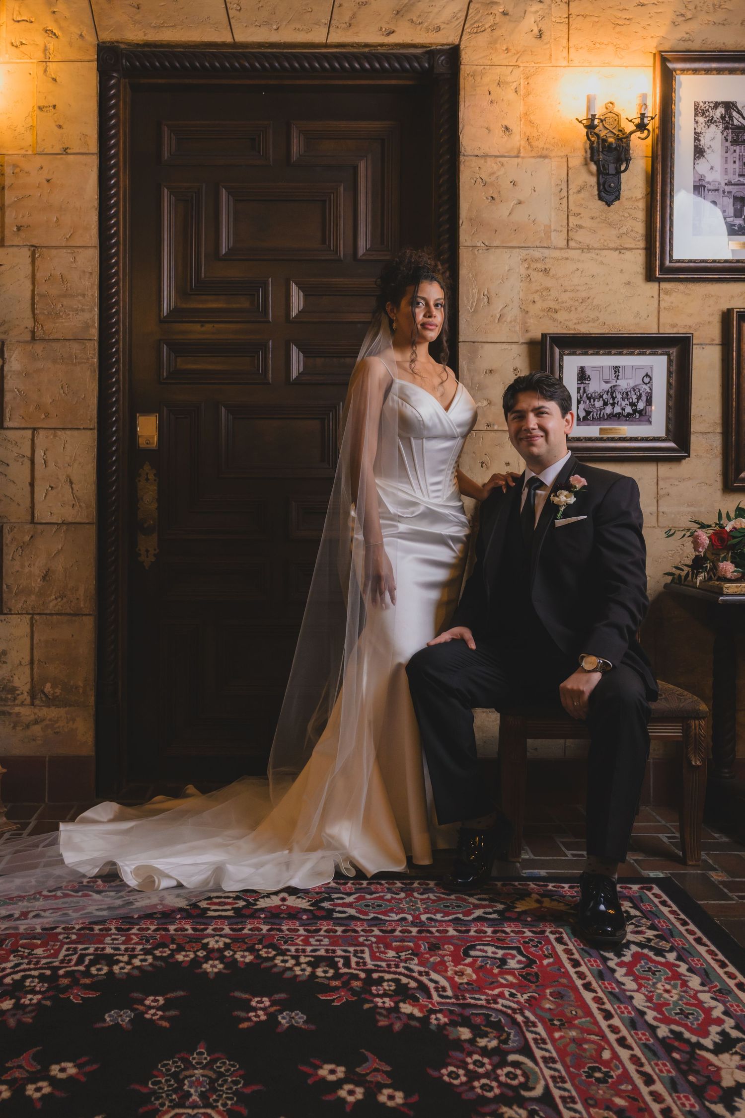 Bride in white gown with veil stands beside seated groom in black suit on ornate rug in elegant stone interior.