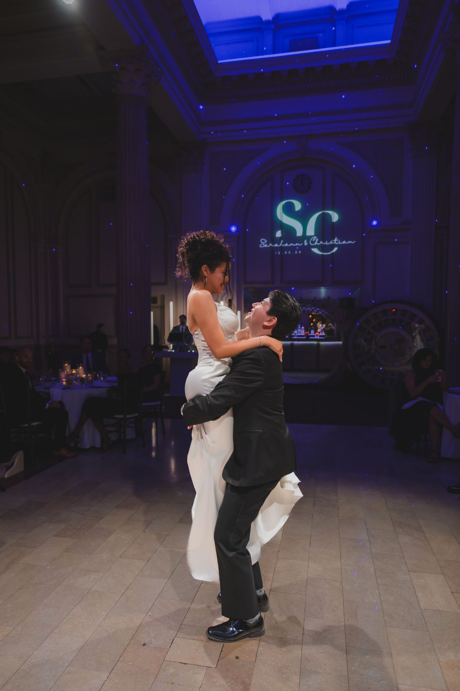 Groom lifts bride during first dance at elegant wedding reception with monogram SC lighting backdrop.
