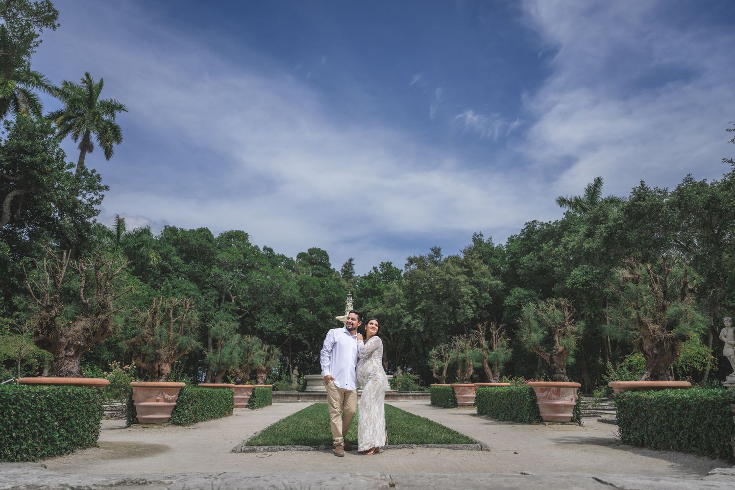 Couple embracing in a lush garden pathway lined with potted plants and palm trees under a bright blue sky.