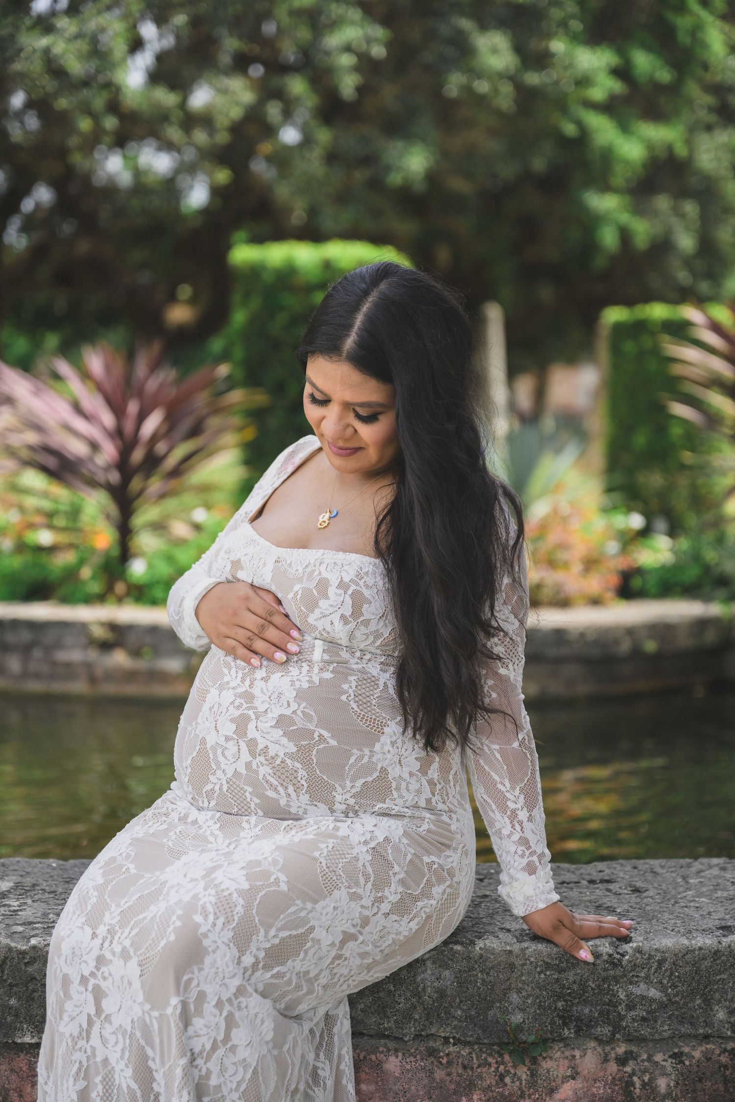 Pregnant woman in white lace gown sitting outdoors by stone wall, cradling baby bump in lush garden setting.