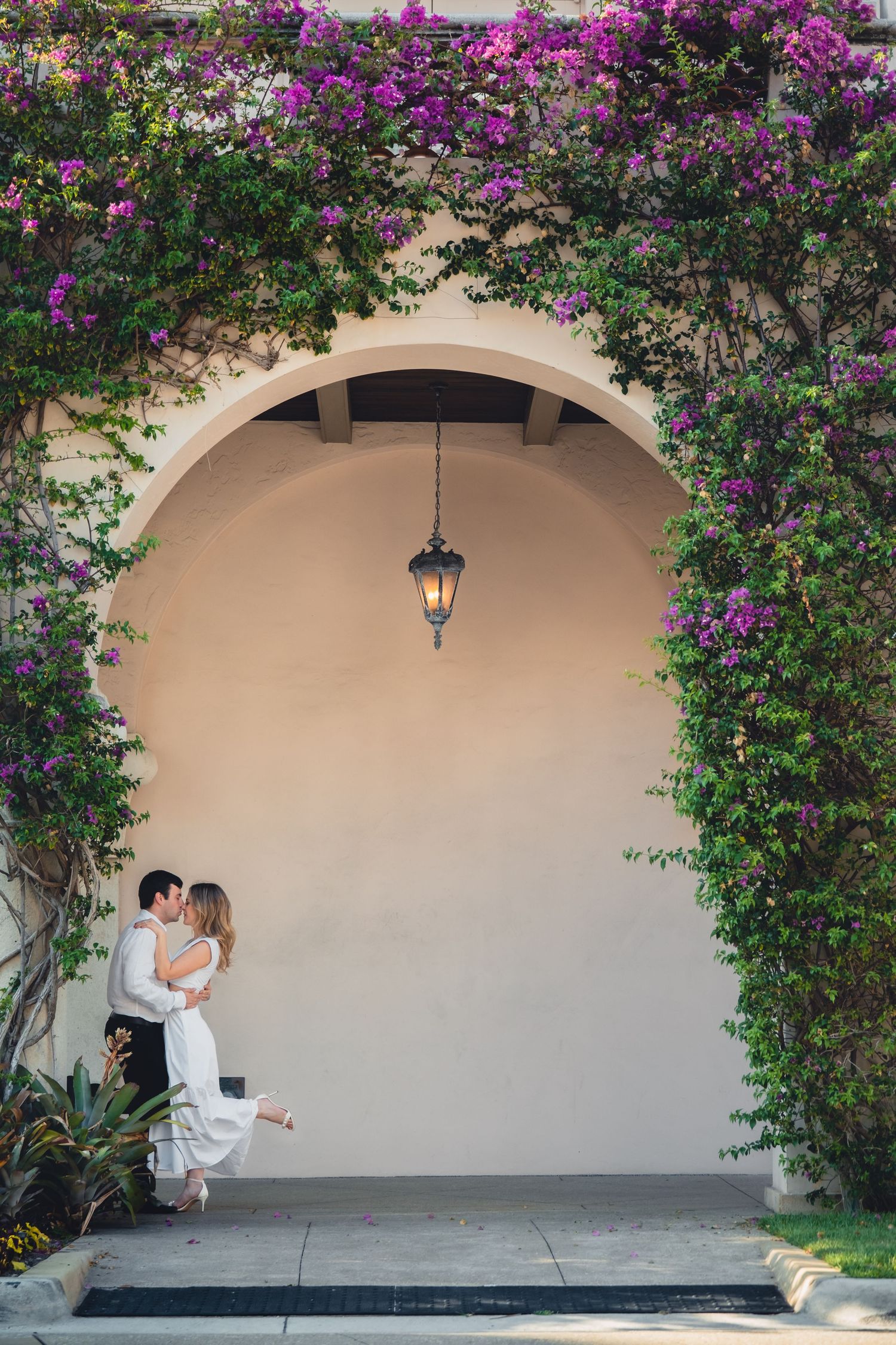 Couple kissing under flower-covered arch with purple bougainvillea and hanging lantern.