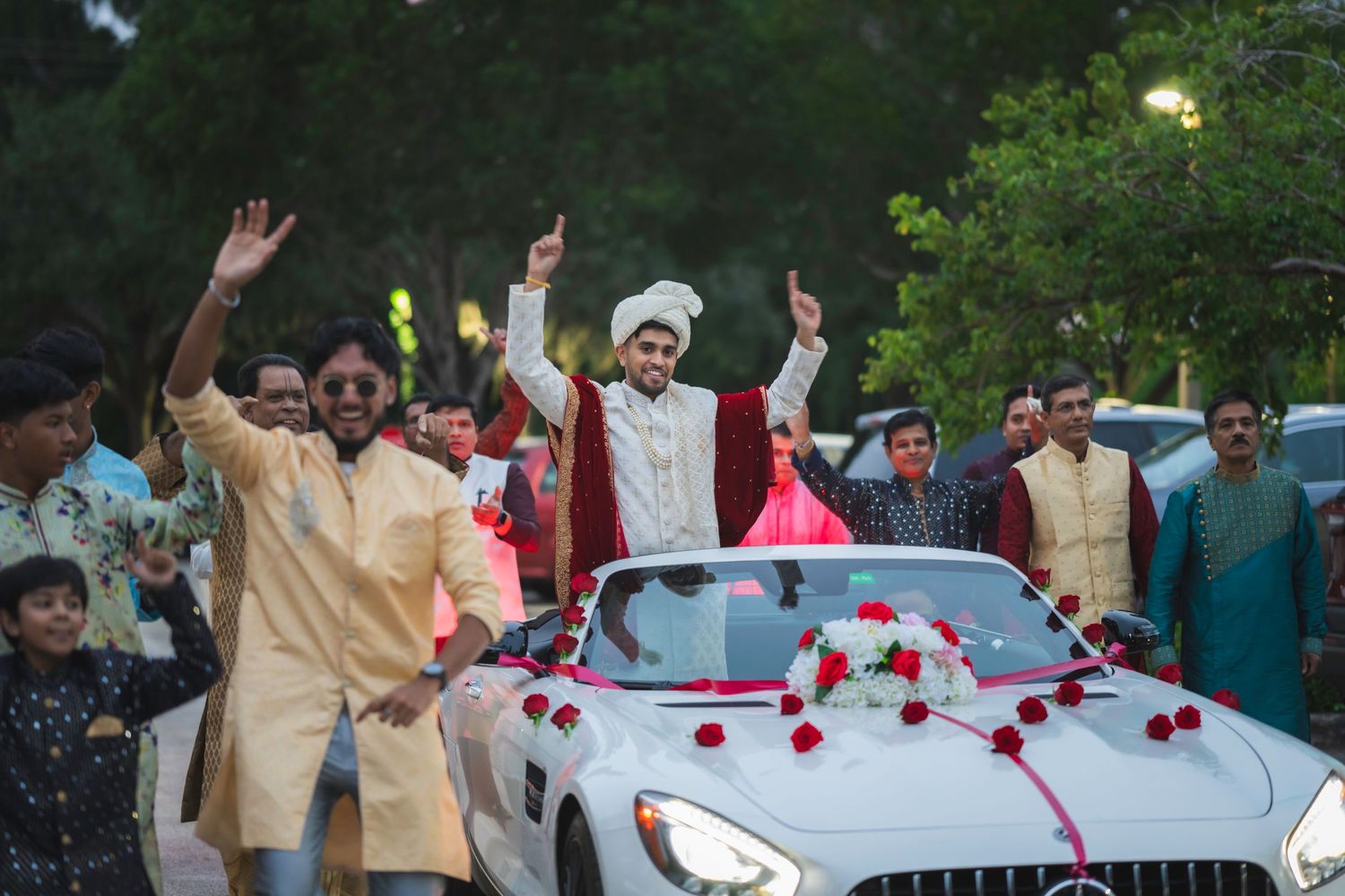 Indian groom in white sherwani stands in decorated Mercedes convertible surrounded by celebrating wedding guests.