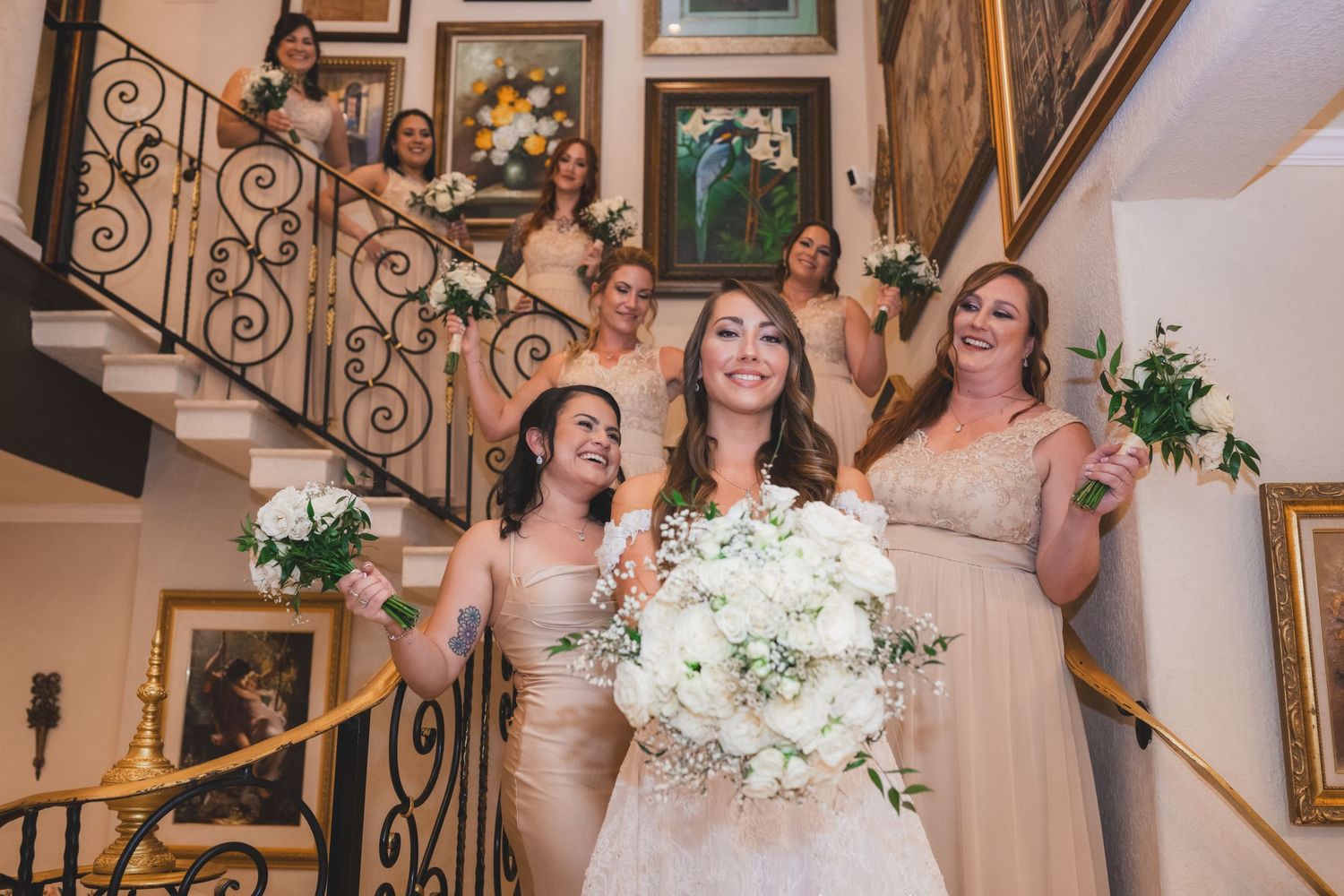 Radiant bride poses on a grand staircase with bridesmaids in champagne gowns holding white bouquets.