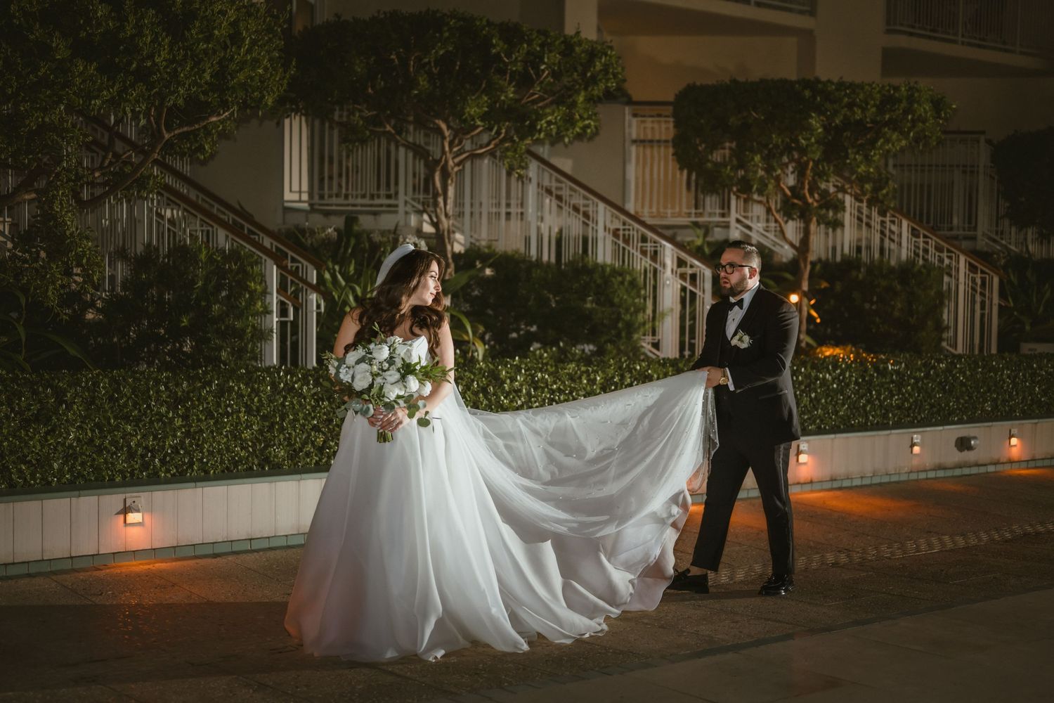 Bride in flowing white gown with groom in tuxedo captured at night outside an elegant wedding venue.