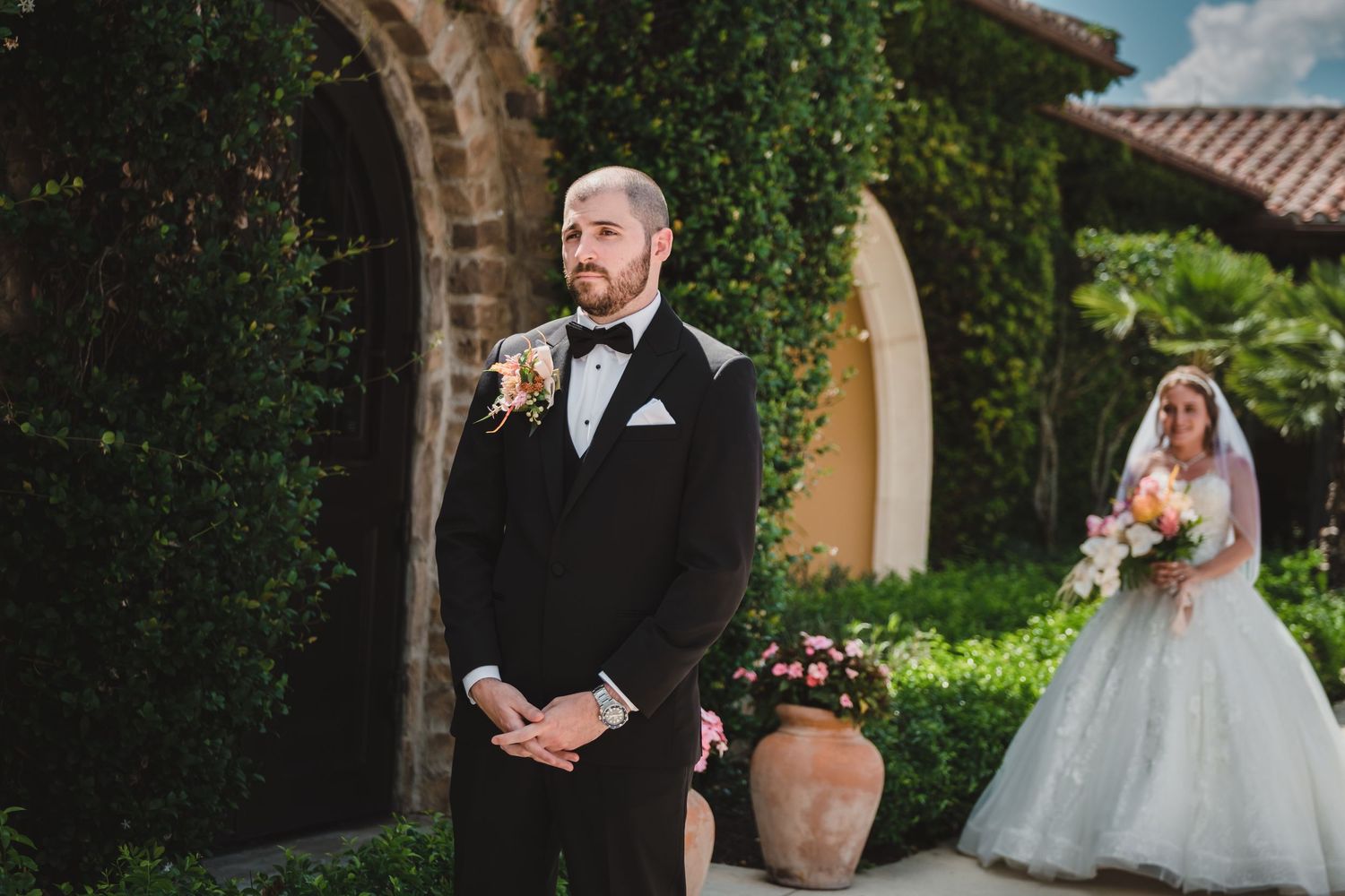 Groom in black tuxedo stands as bride in ball gown approaches during outdoor first look at villa.