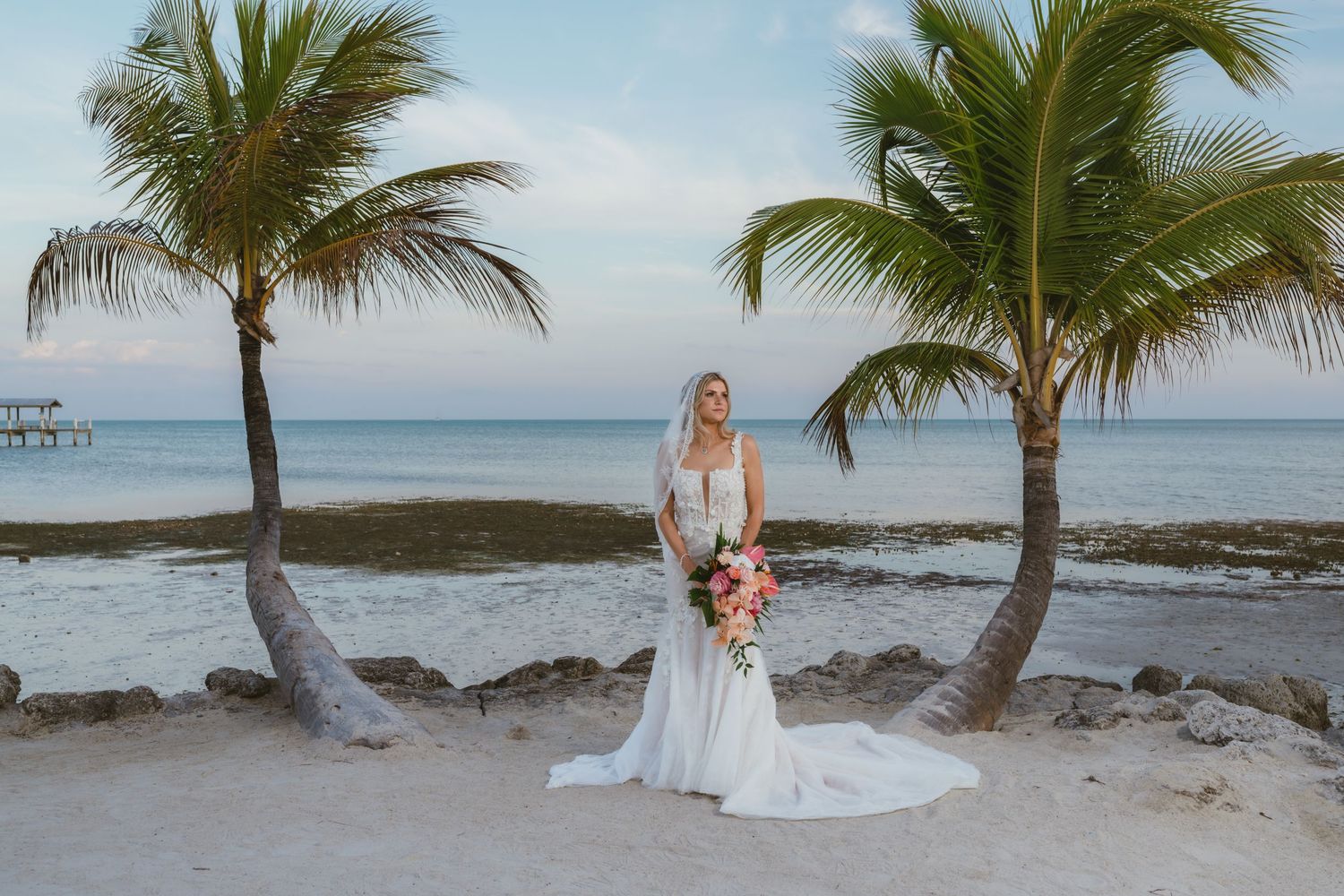Bride in fitted white gown with veil stands between two palm trees on tropical beach.