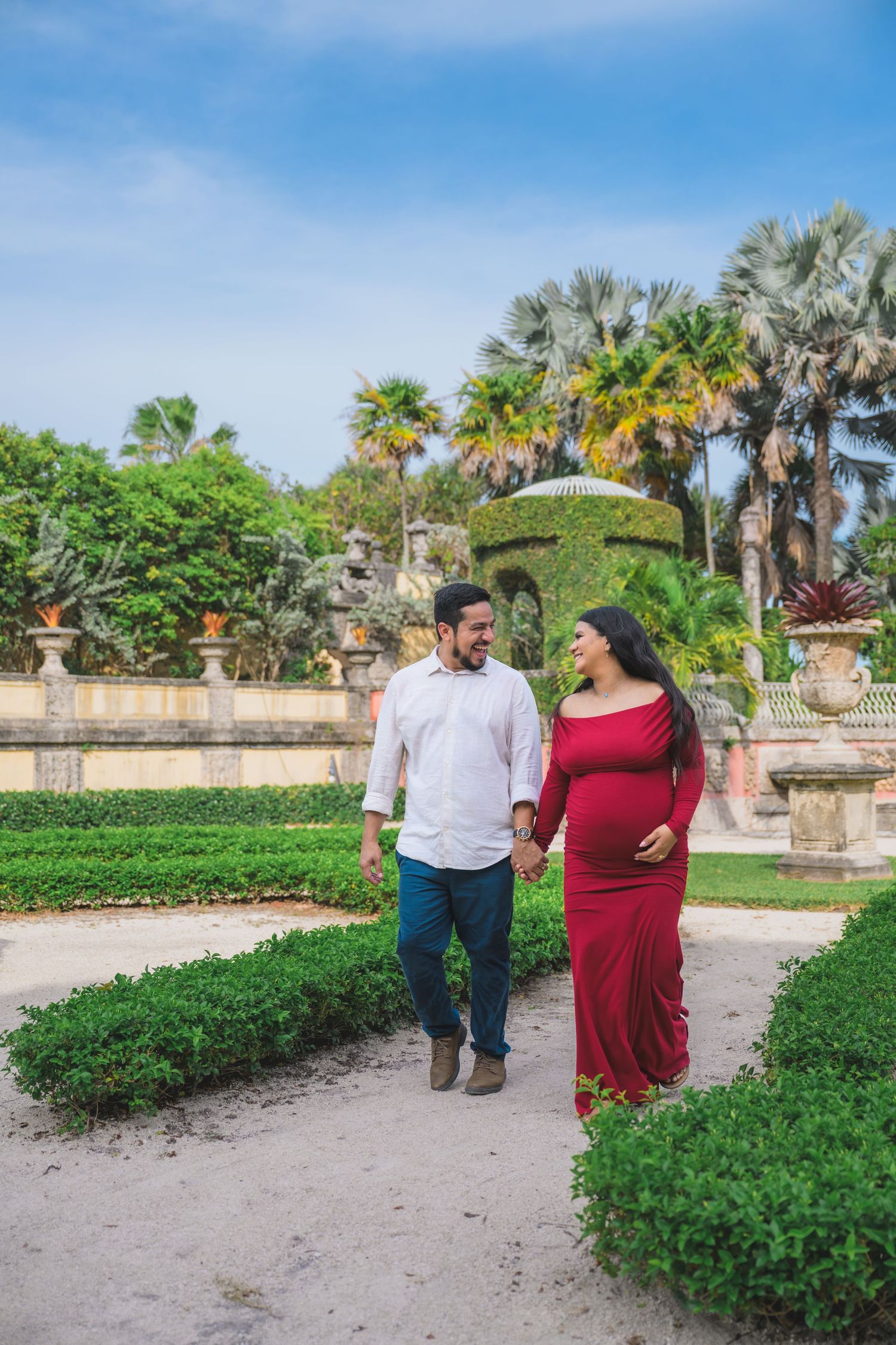 Expecting couple holding hands walking through lush tropical garden, woman in red maternity gown.