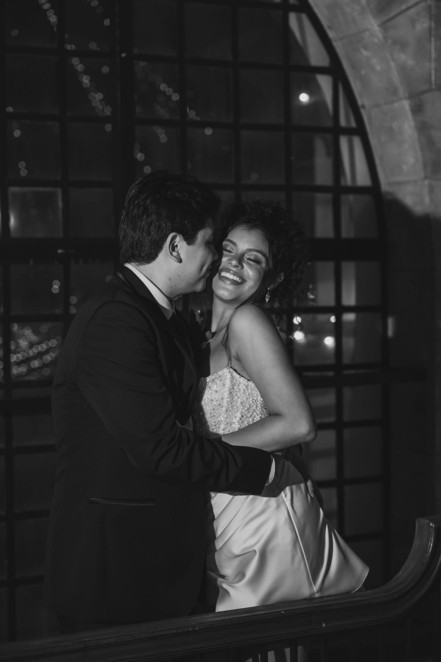 Black and white photo of a smiling bride and groom embracing near a large arched window at night.