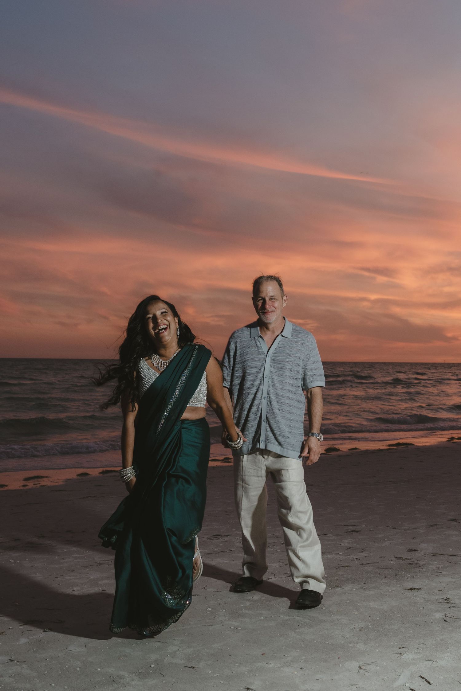 Couple holding hands laughing on beach at sunset, woman in teal saree and man in casual attire.