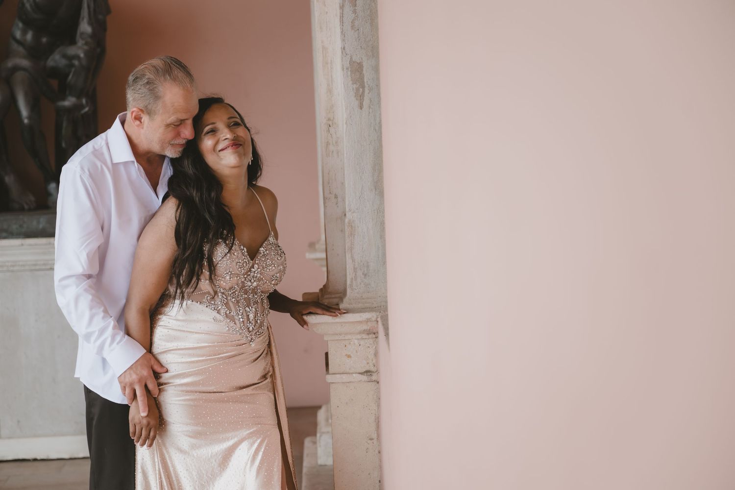 Couple in elegant wedding attire embracing near a pink wall, bride in beaded gown smiling up at groom in white shirt.