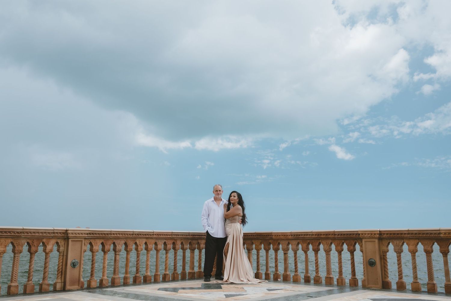 Couple posing on ornate stone balcony terrace with ocean view and dramatic cloudy sky backdrop.