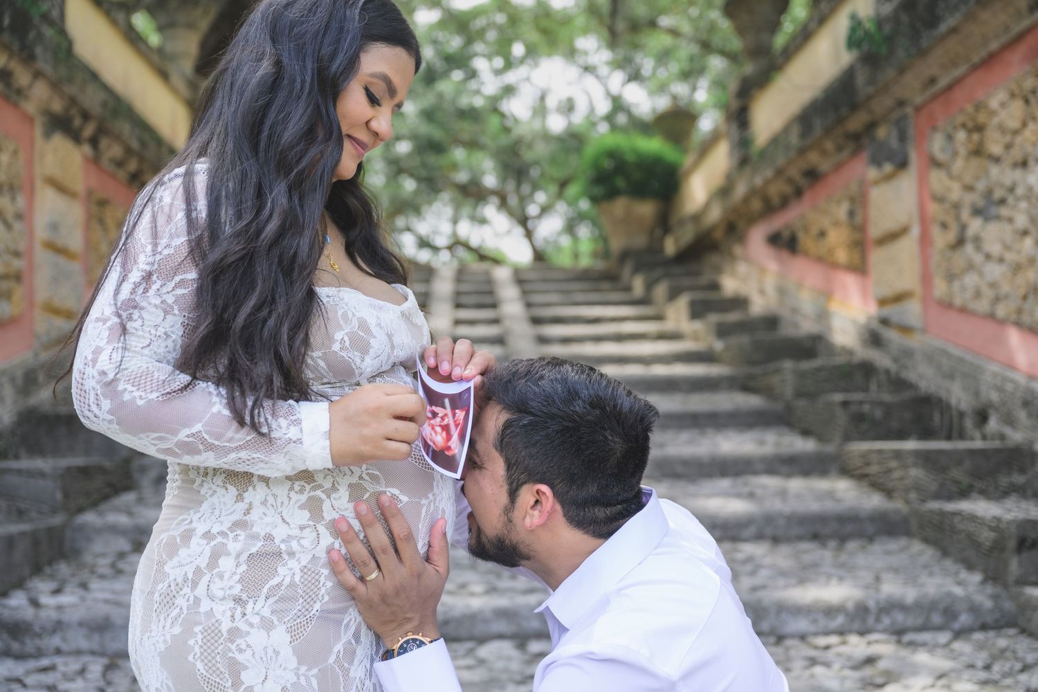 Pregnant woman in white lace dress with man kneeling kissing her belly holding baby shoes outdoors on stone steps.