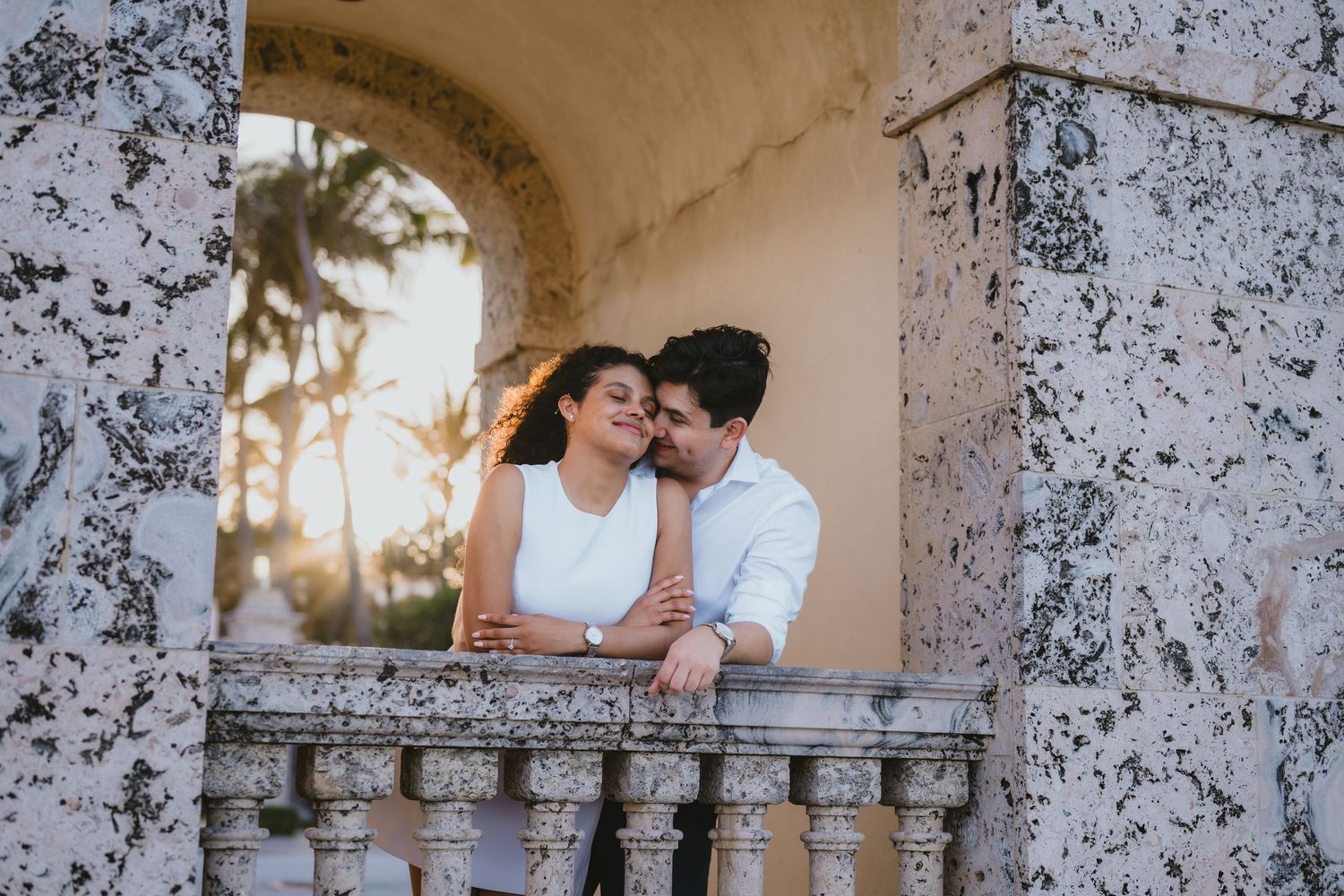 Couple sharing tender moment on ornate stone balcony at golden sunset.
