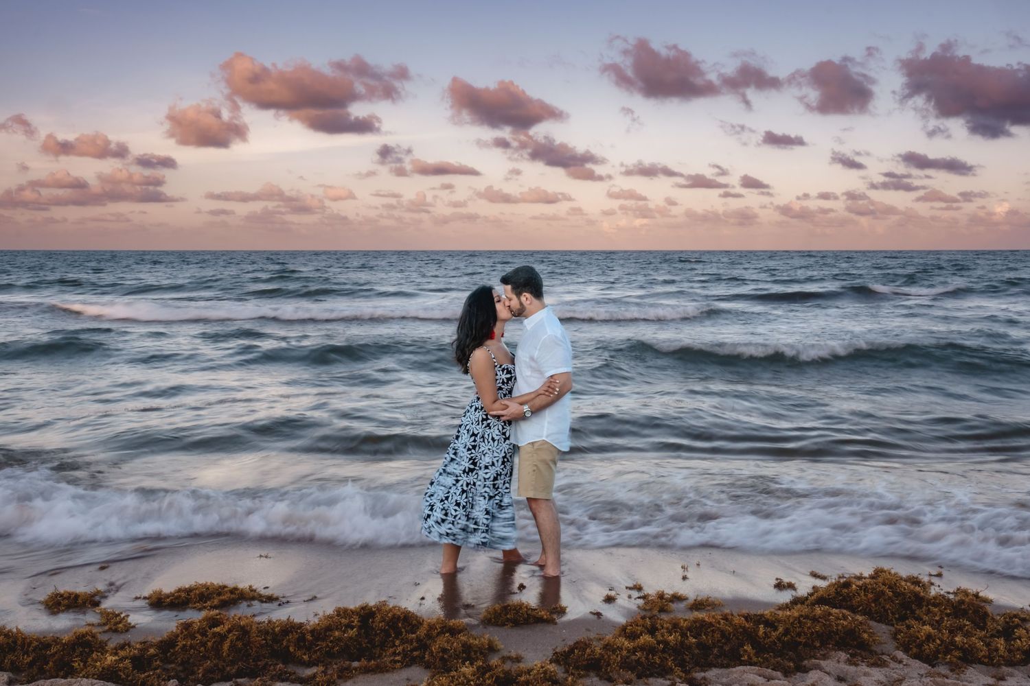 Couple sharing a romantic kiss on a sandy beach at sunset with colorful sky.