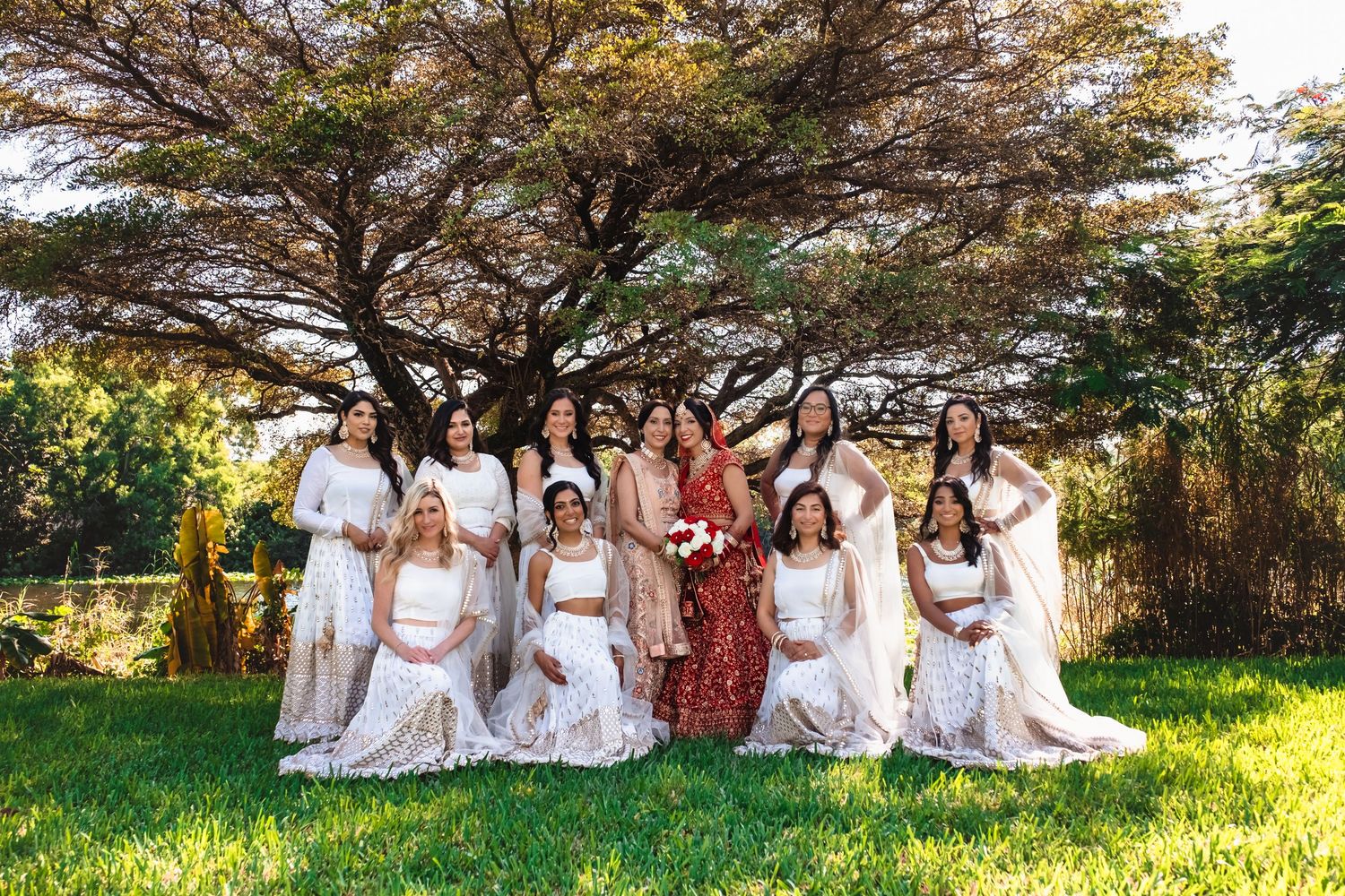 Indian bride in red lehenga with bridesmaids in white posing under a large tree outdoors.