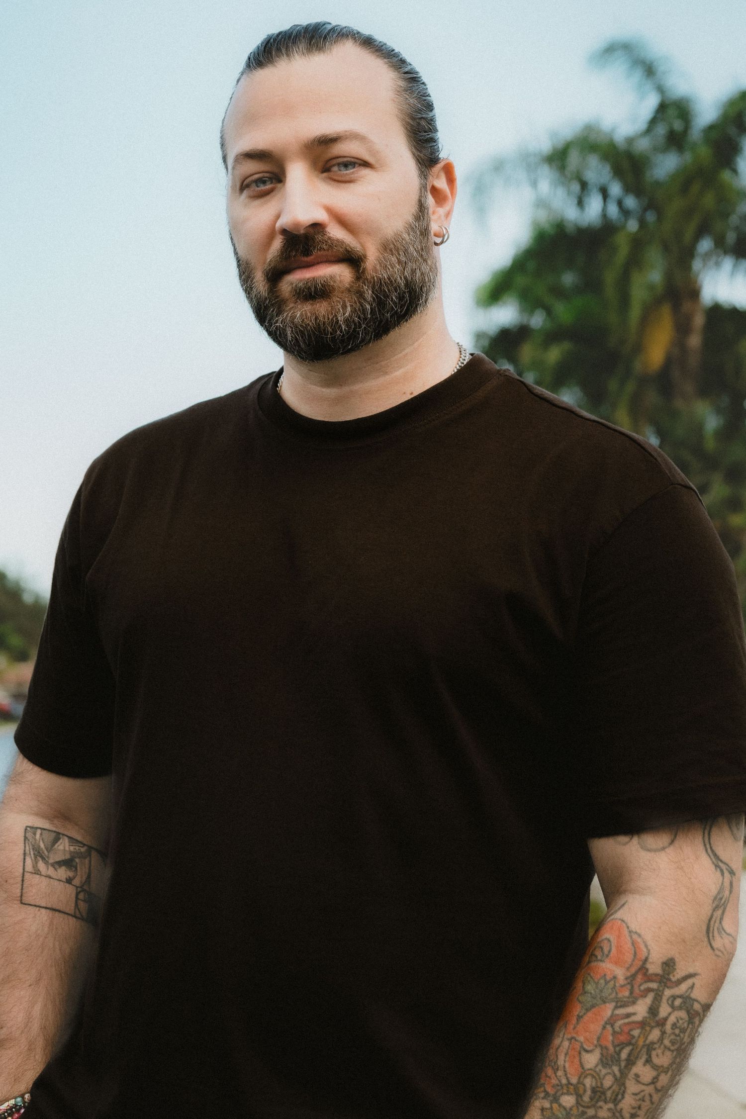Bearded tattooed man in black t-shirt poses outdoors with palm trees behind him.