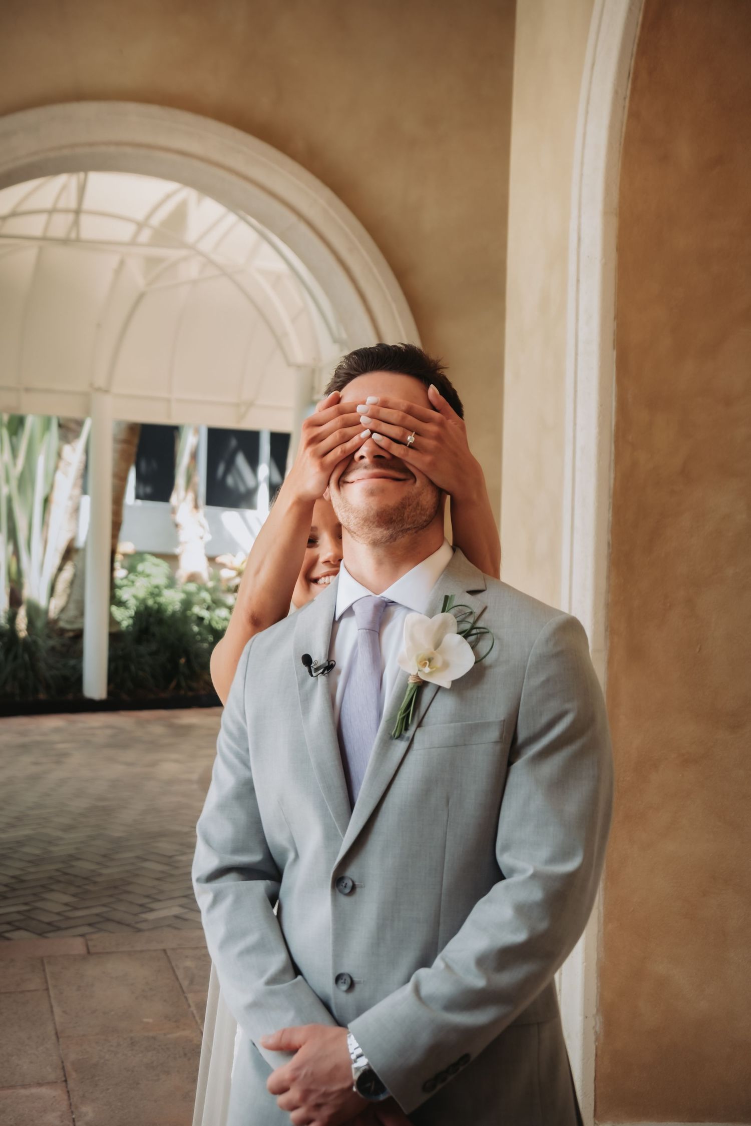 Bride covers grooms eyes with hands during playful first look moment under arched walkway.