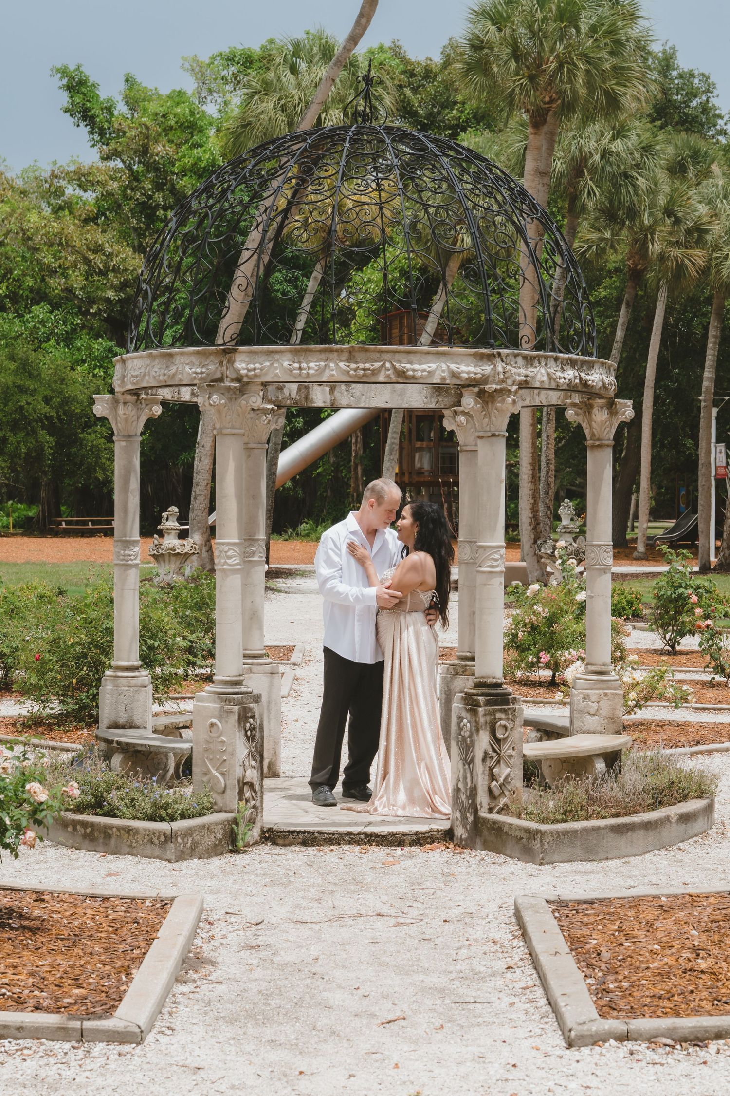 Couple embracing under an ornate stone gazebo with wrought iron dome in a lush garden setting.
