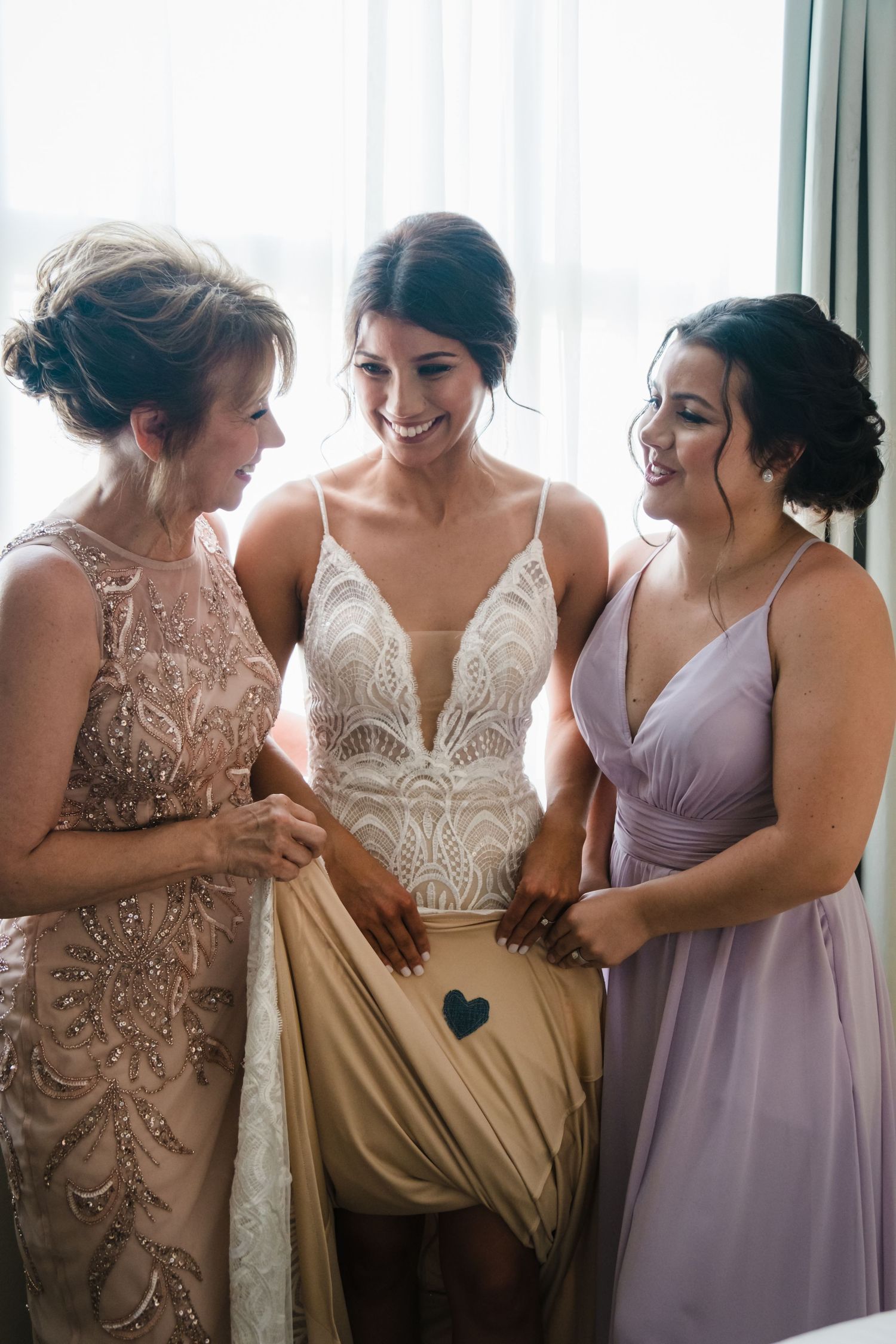 Bride in a lace gown smiles joyfully while flanked by her mother and bridesmaid near a window.