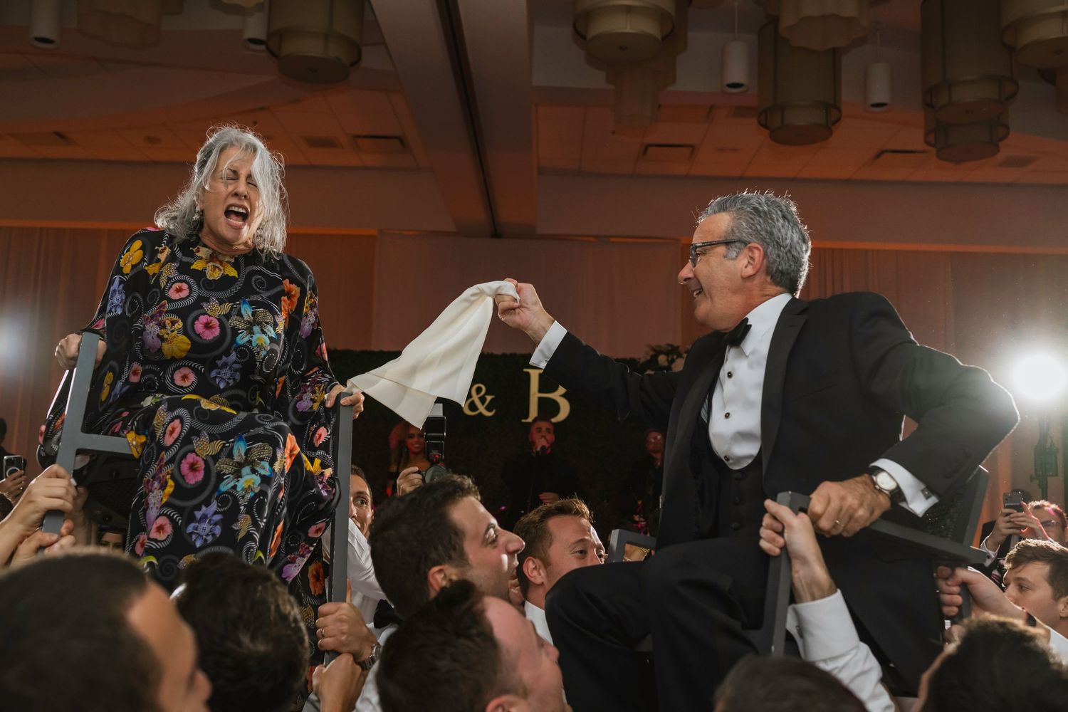Older couple lifted by crowd on chairs during festive Jewish wedding hora dance celebration.