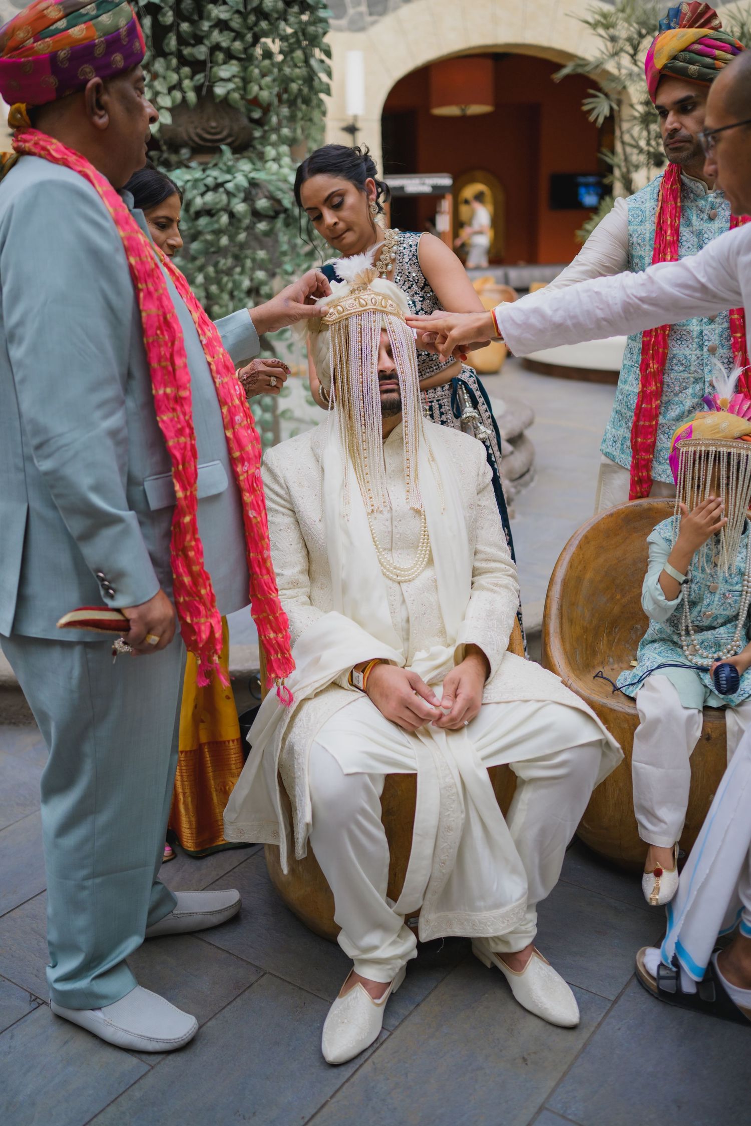 Groom in white sherwani receives traditional sehra headpiece during Indian wedding preparation ceremony.