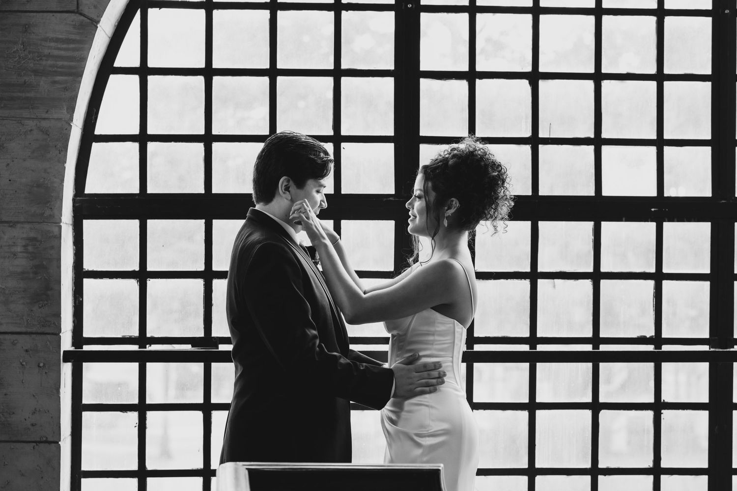 Black and white photo of bride and groom sharing tender first look moment by large window.