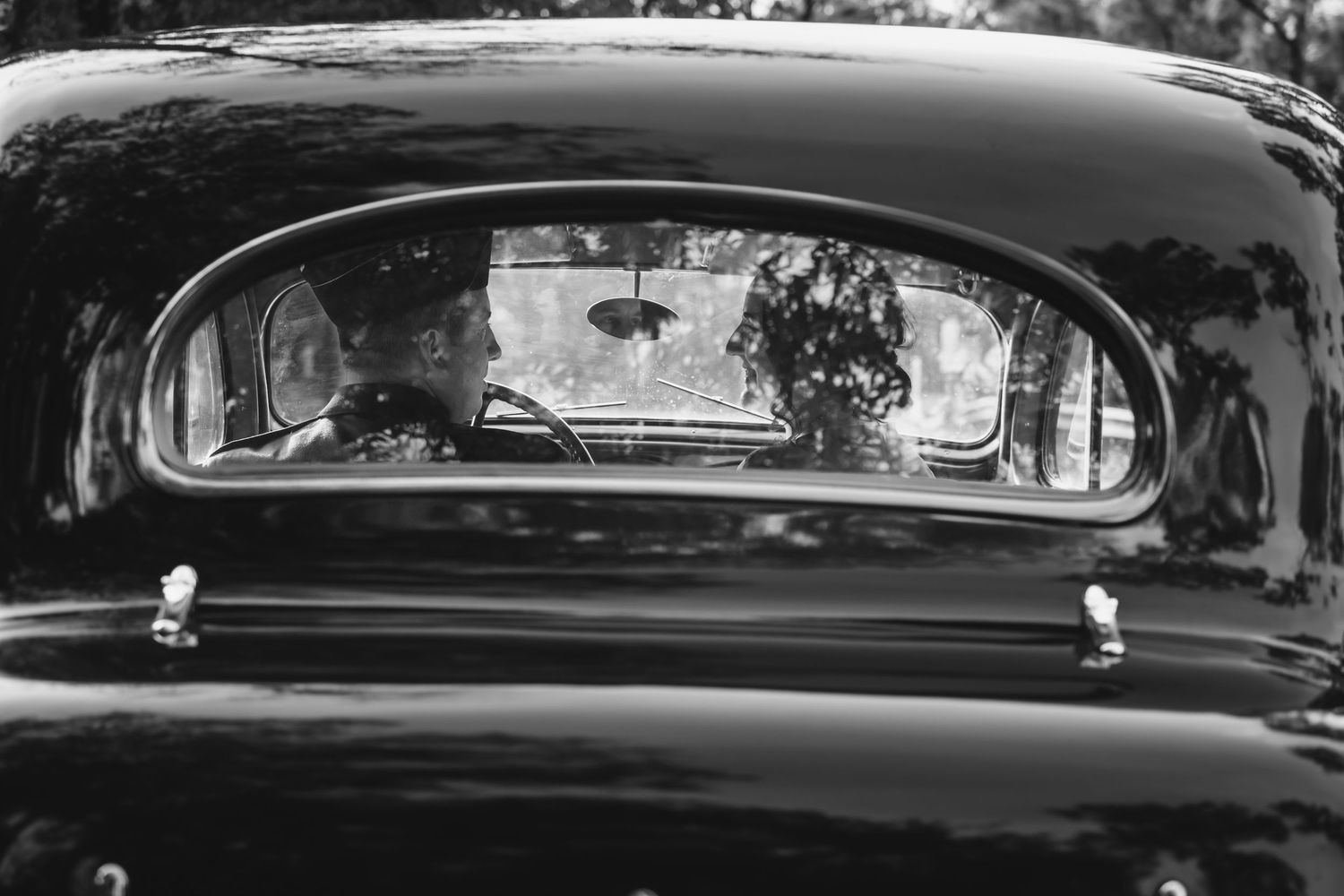 Black and white reflection of bride and groom visible through rear window of a vintage classic car.