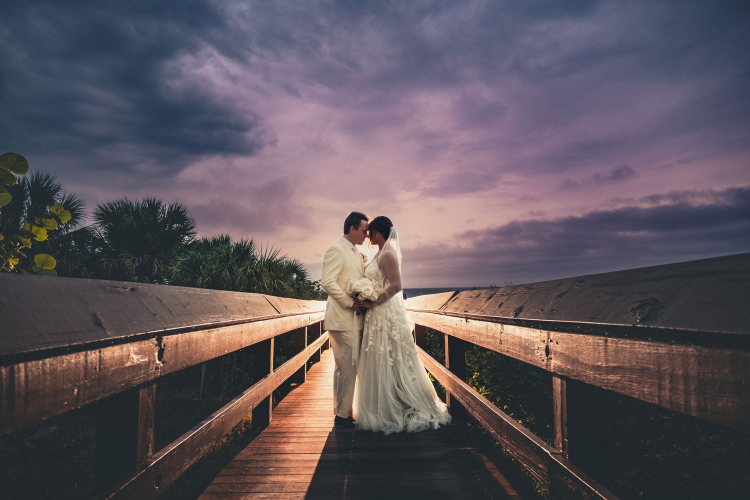 Bride and groom share a romantic moment on a wooden bridge at sunset with dramatic purple clouds.