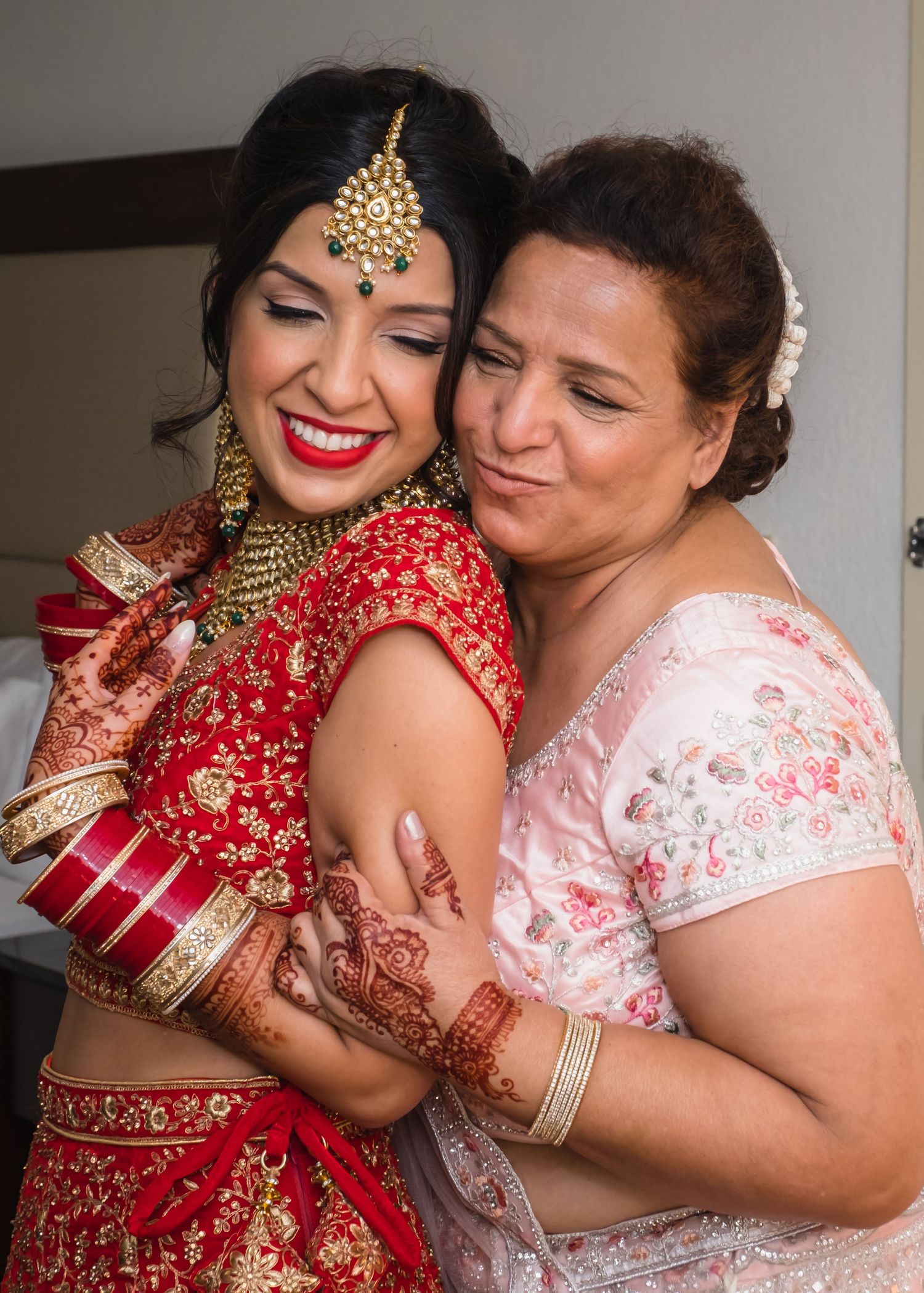 Indian bride in red lehenga with mehndi embracing her mother during wedding preparations.