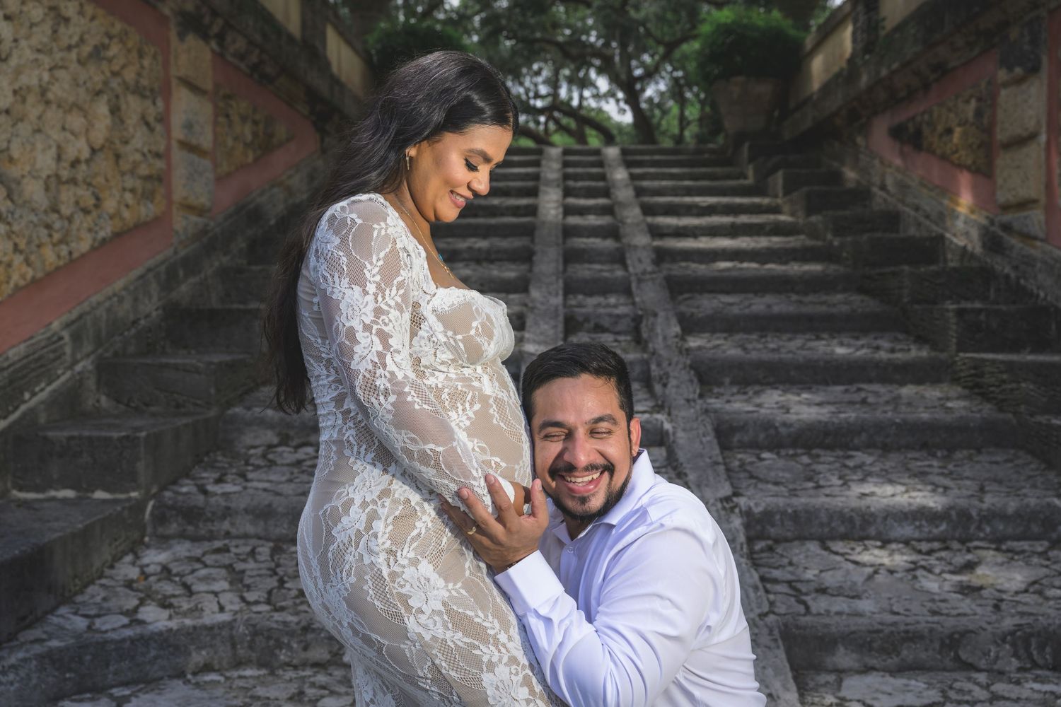 Pregnant woman in lace dress with partner kneeling beside her on stone steps during maternity photo shoot.