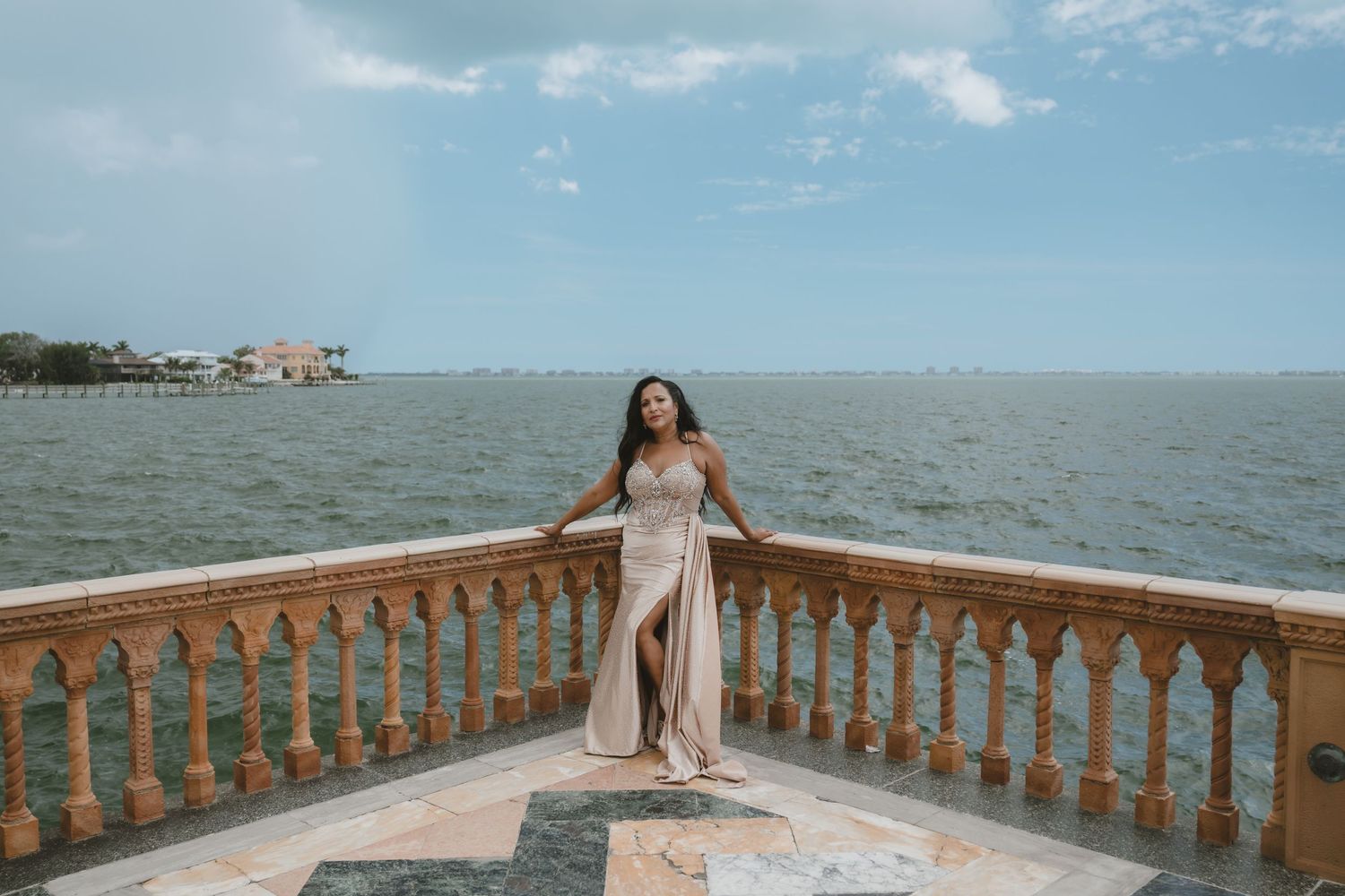Woman in elegant satin gown poses on a stone balcony overlooking a serene bay under cloudy skies.