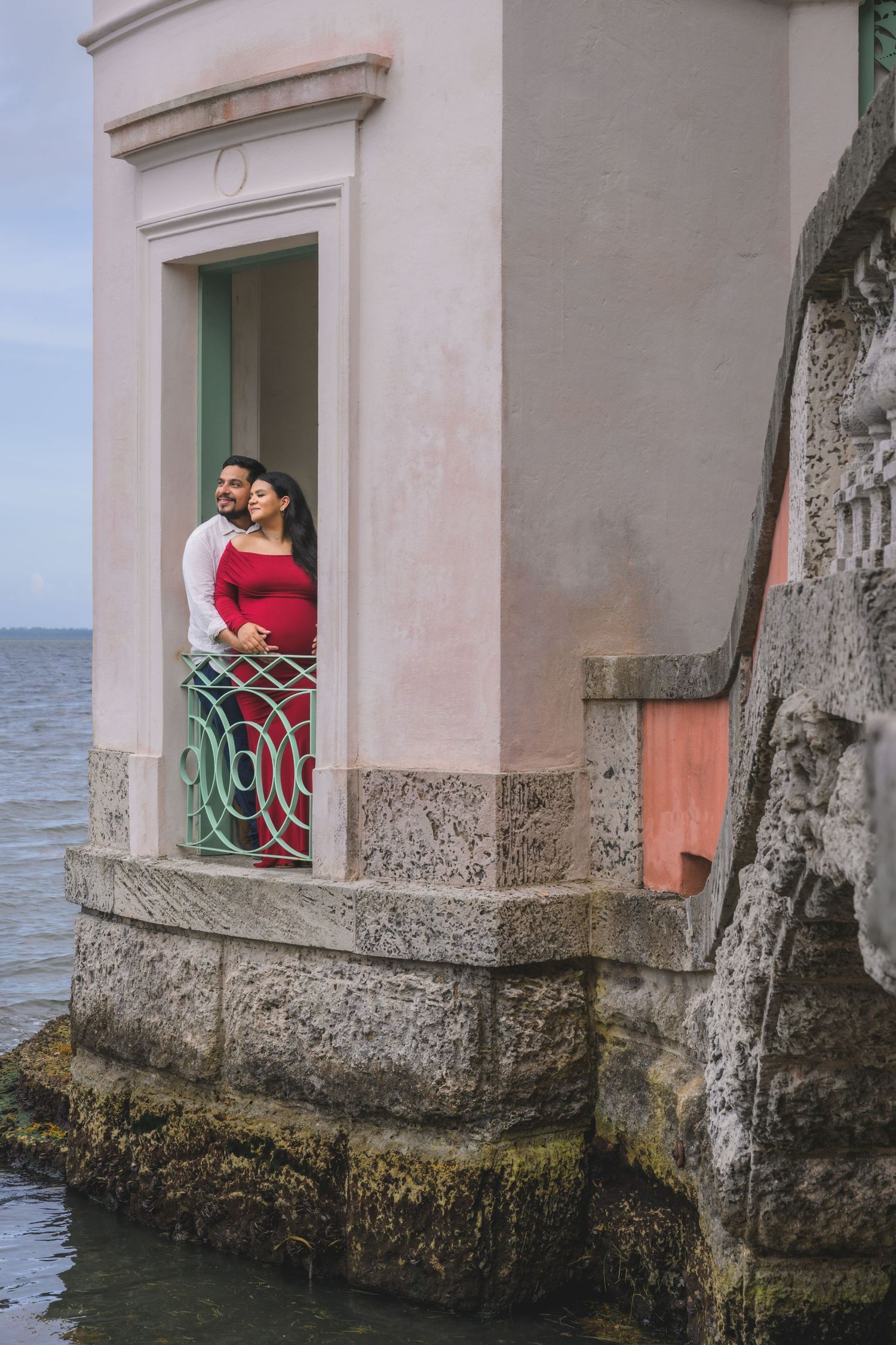 Couple standing on waterfront balcony of pink historic building overlooking calm ocean waters.