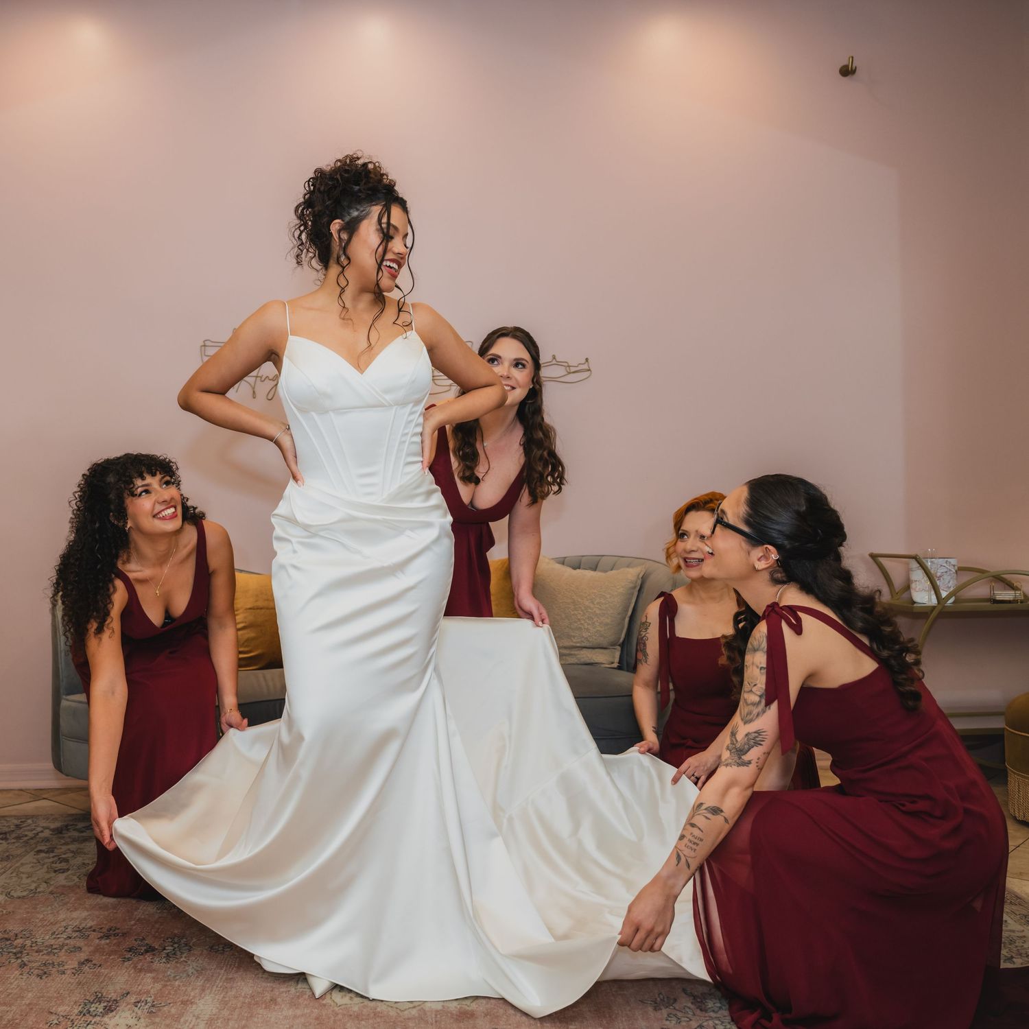 Bride in white gown laughs as bridesmaids in burgundy dresses help arrange her train before the wedding ceremony.