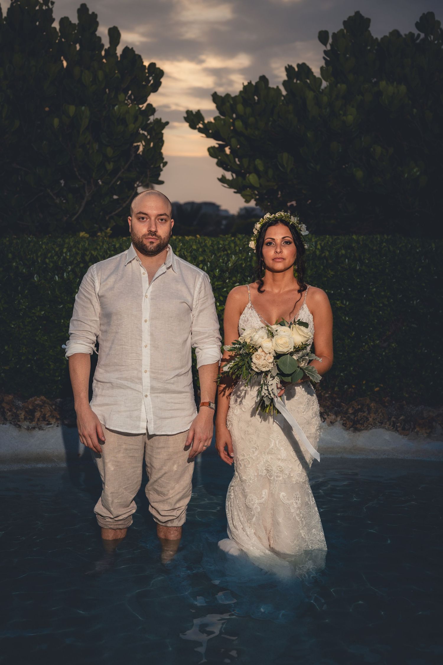 Bride and groom standing in a pool at sunset, bride holding white floral bouquet.