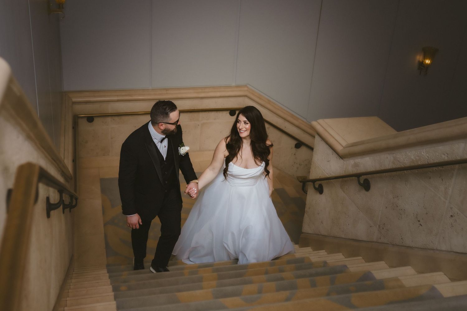 Bride in tulle gown and groom walk hand in hand up marble stairs at elegant wedding venue.