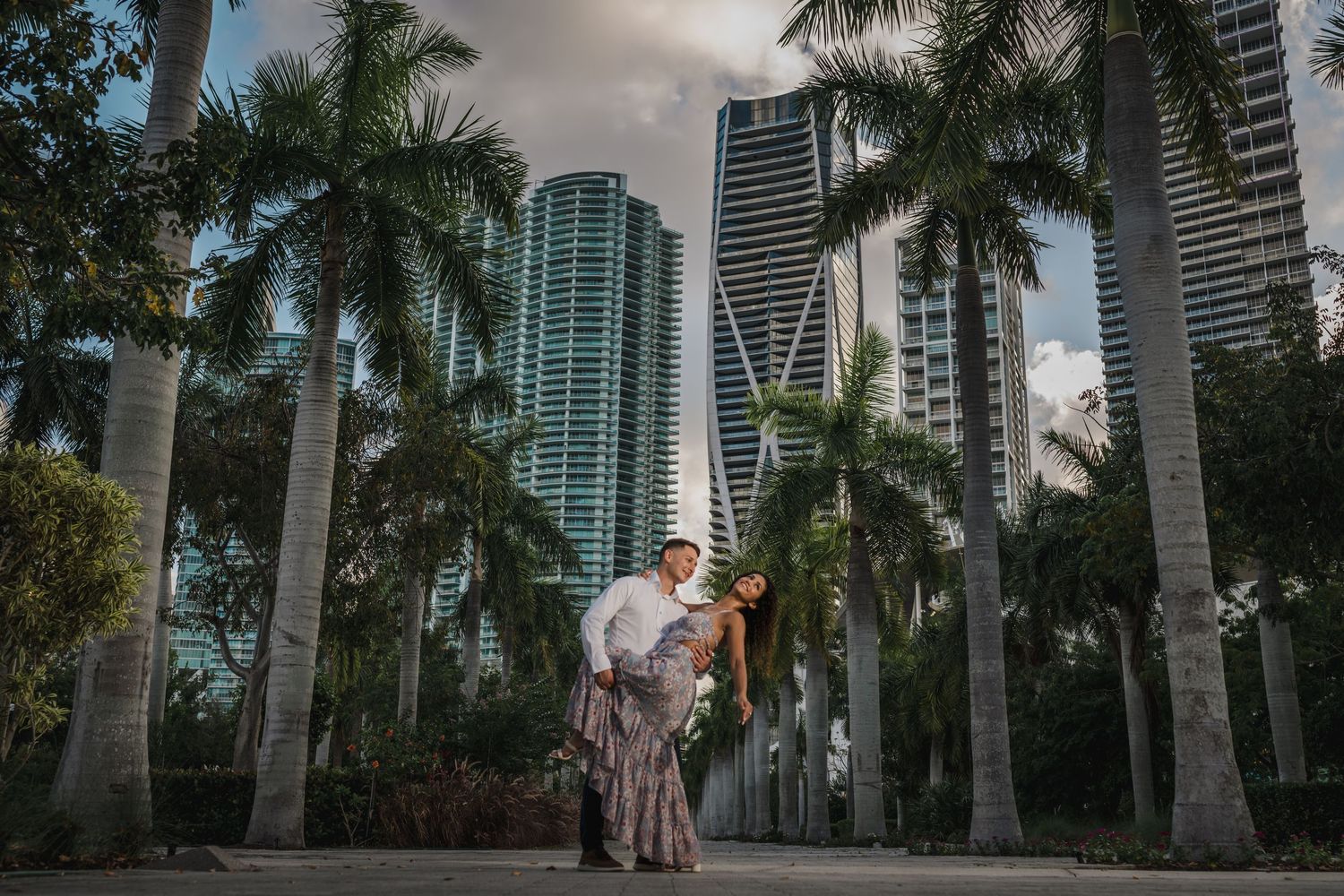 Couple poses romantically among tall palm trees with Miami city skyscrapers in the background.