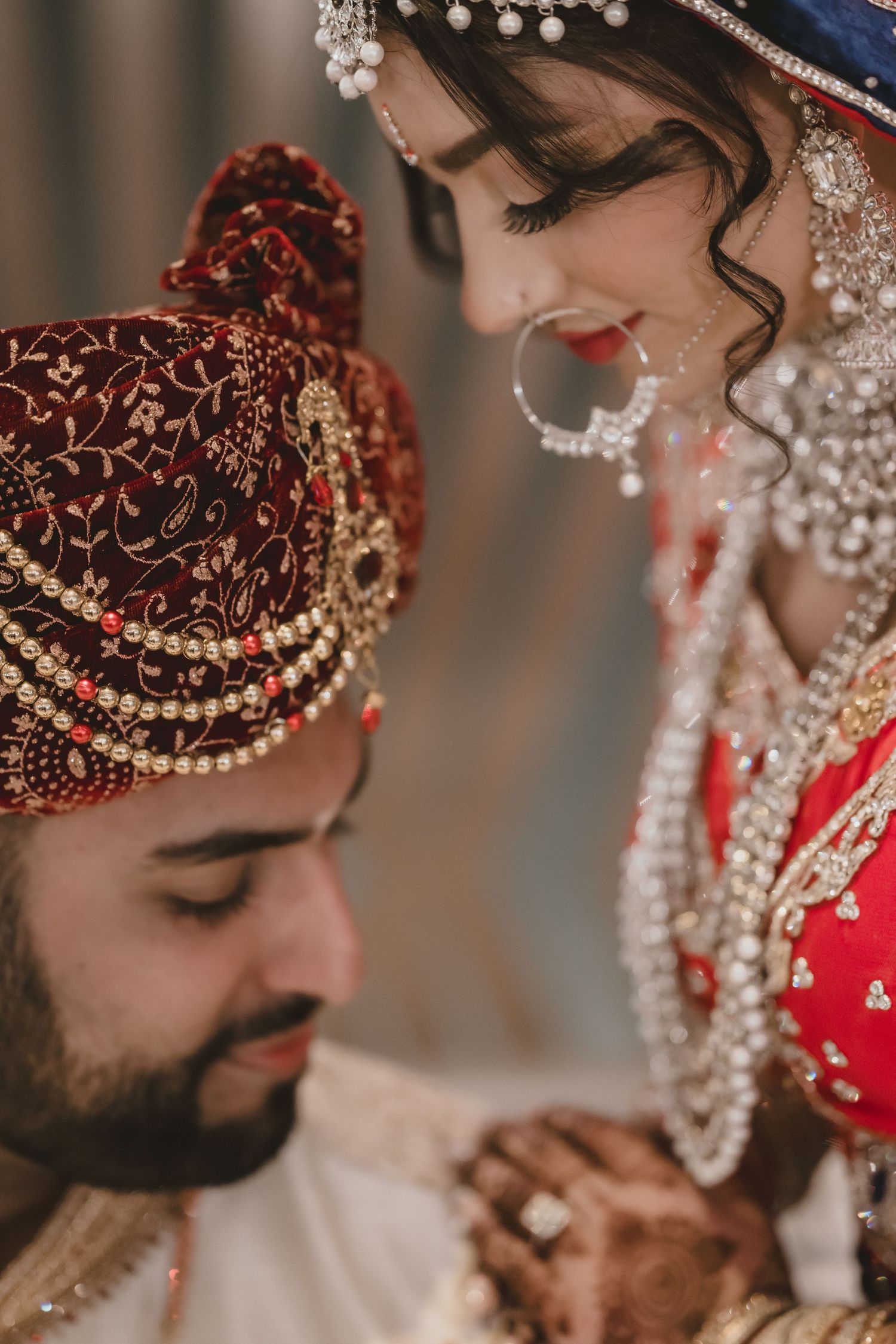 Indian bride in red lehenga adorned with jewelry gazing down at groom during wedding ceremony.