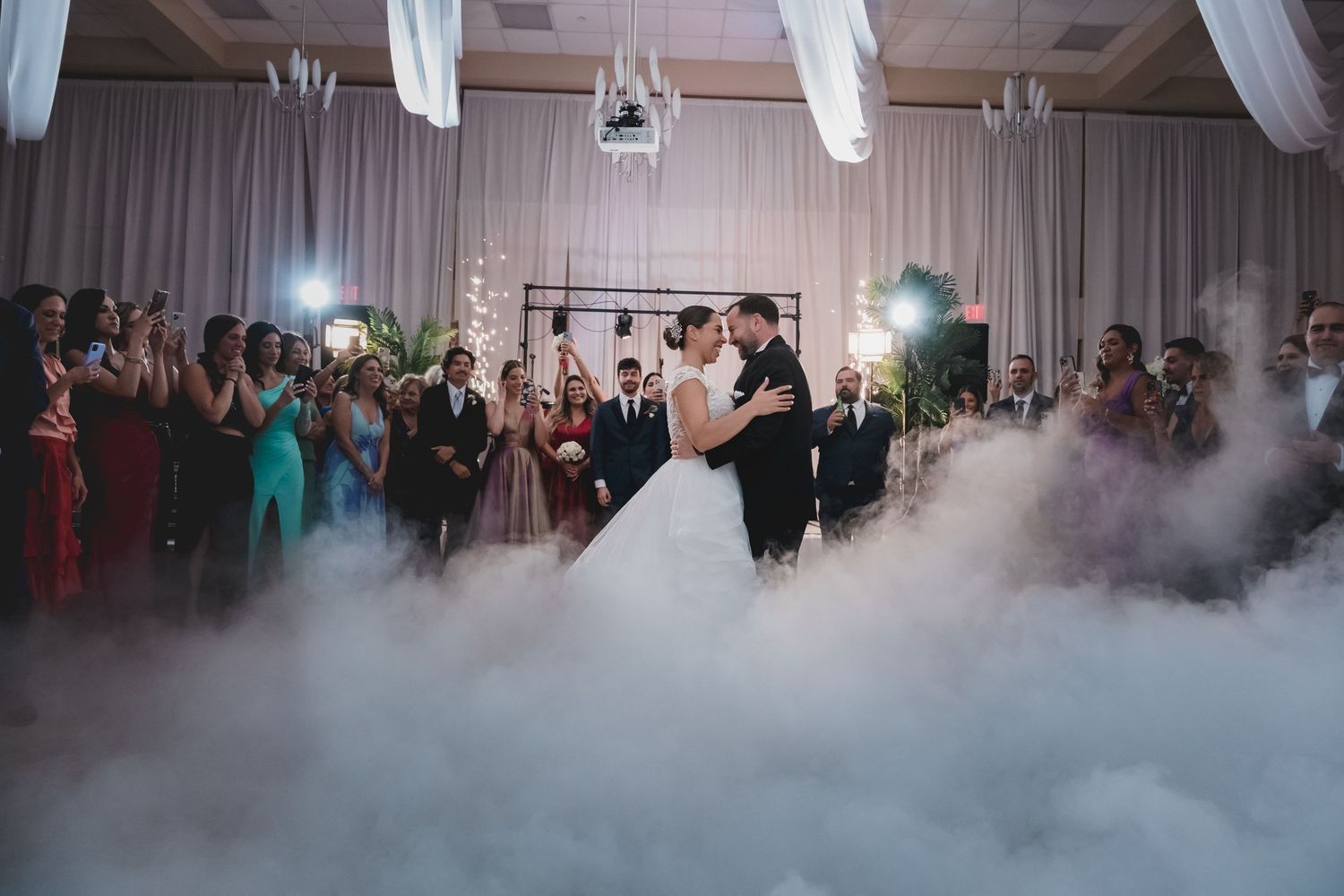 Bride and groom share first dance surrounded by dry ice fog and guests at elegant wedding reception.