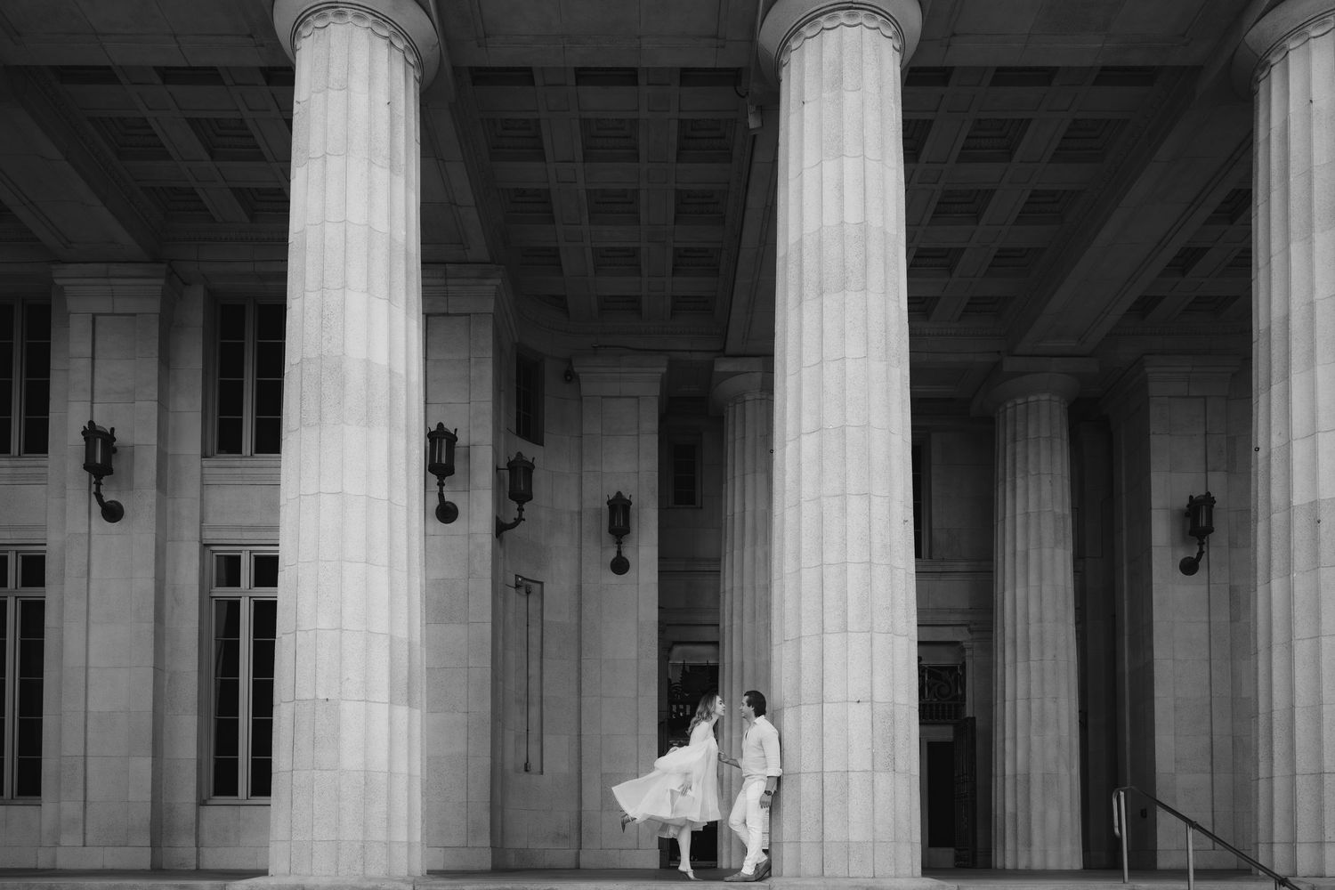 Black and white photo of a couple in wedding attire standing between grand columns of a neoclassical building.