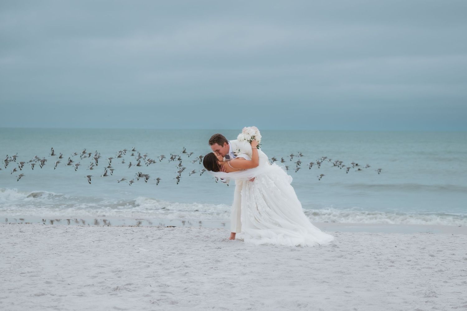 Groom dipping bride in white dress on a beach with birds flying over the ocean waves.
