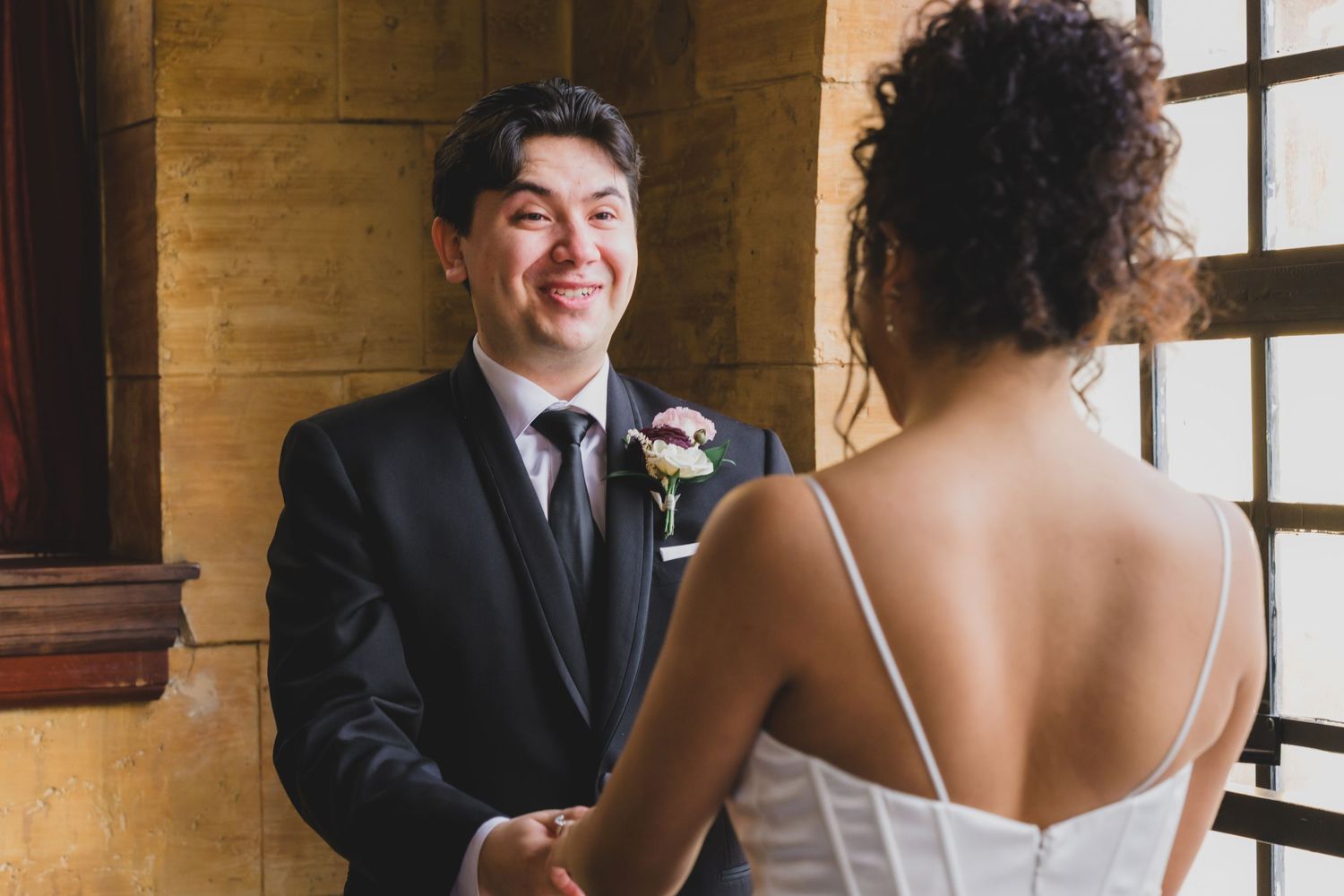 Groom in black suit smiling warmly at bride during intimate first look inside historic venue.