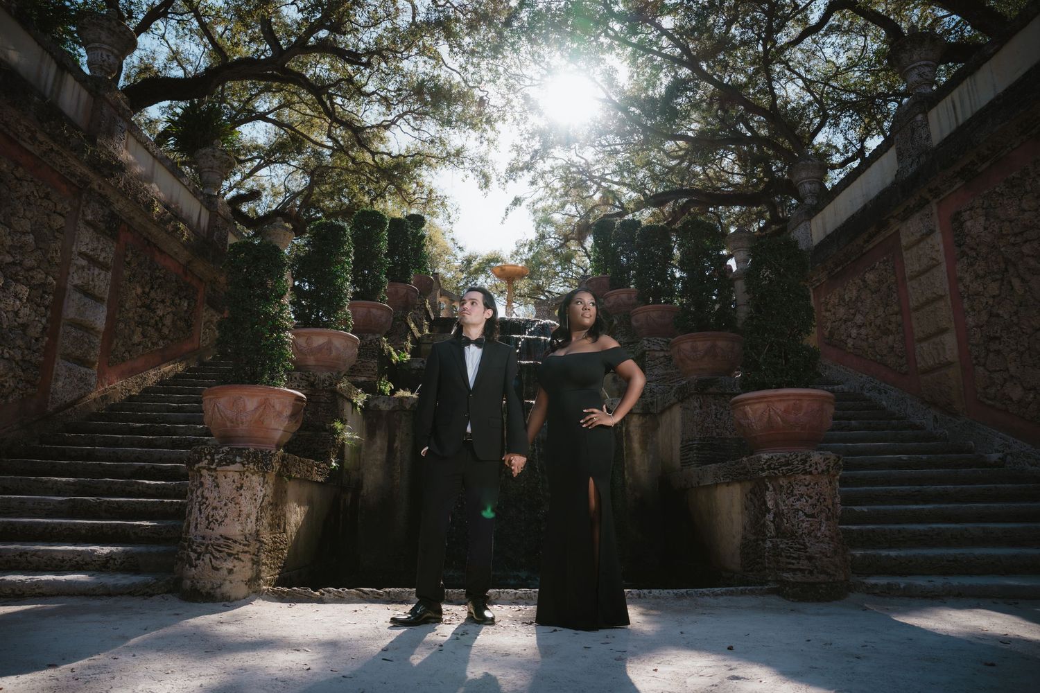 Couple in elegant black formal attire posing on stone steps surrounded by lush greenery.