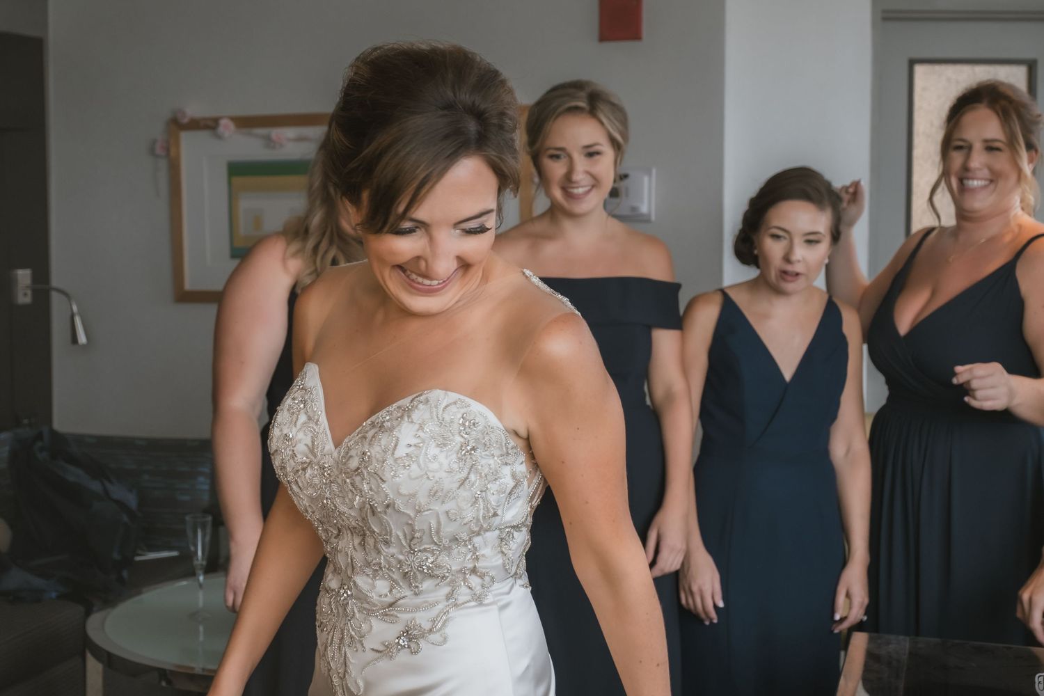 Bride in beaded strapless gown smiles as bridesmaids in navy dresses help her get ready.